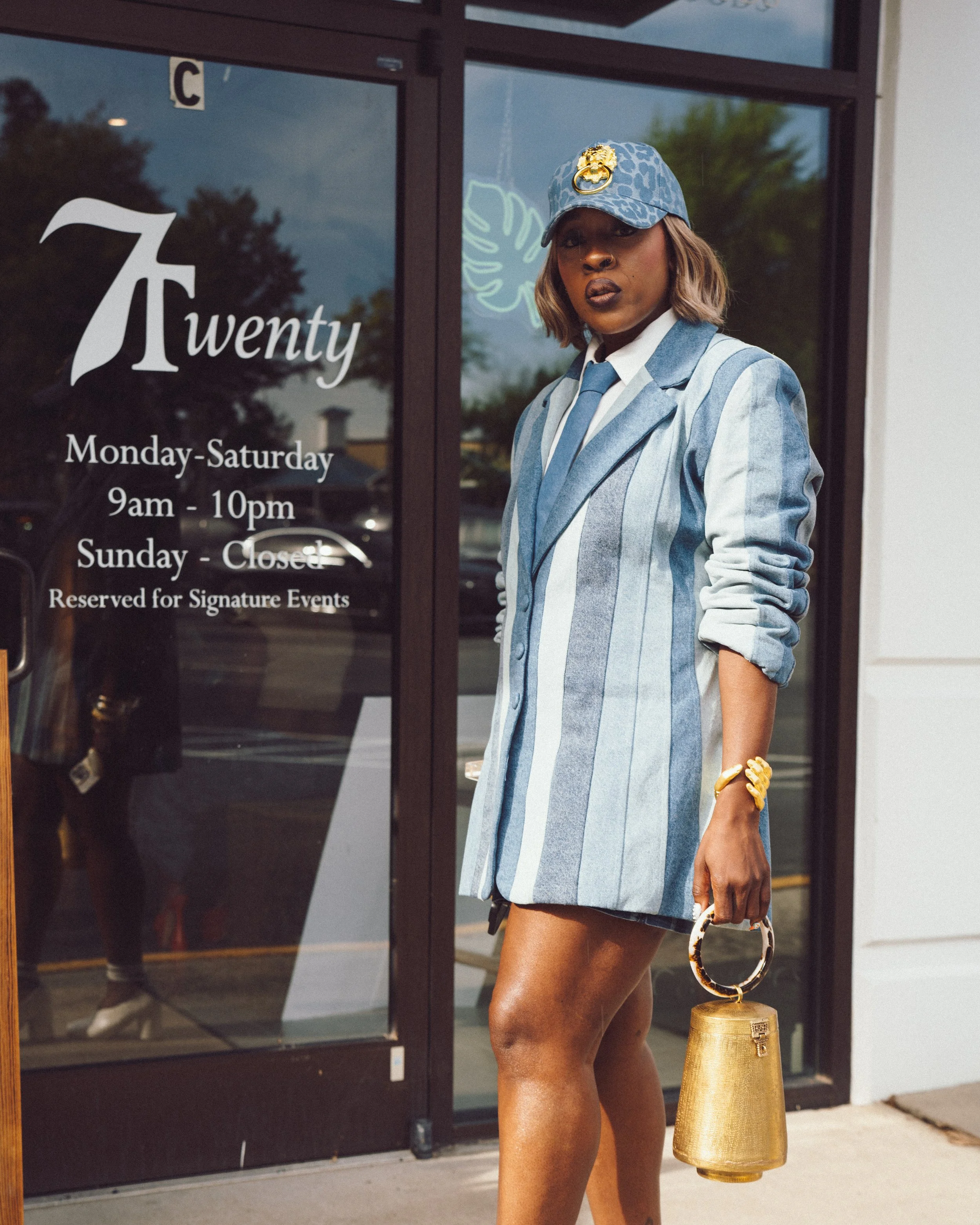 A woman stands outside a storefront wearing a blue and white striped blazer, blue cap with a gold lion emblem, matching blue tie, and holding a gold handbag. The store sign shows hours of operation.