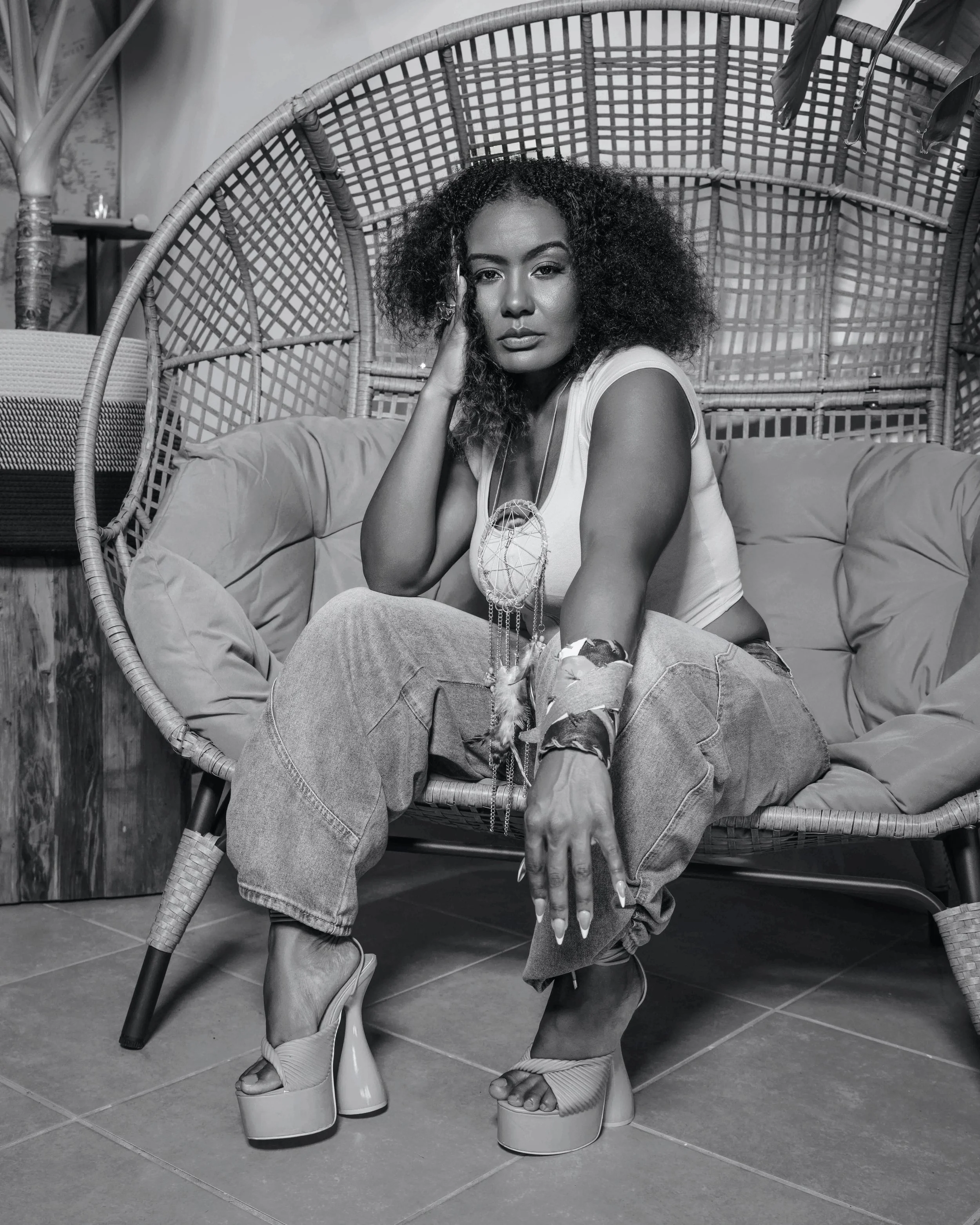 A woman with curly hair sitting in a round wicker chair with a high back. She is wearing a sleeveless shirt and ripped jeans, posed with one hand touching her face and the other resting on her knee. She is wearing platform shoes and various jewelry, 
