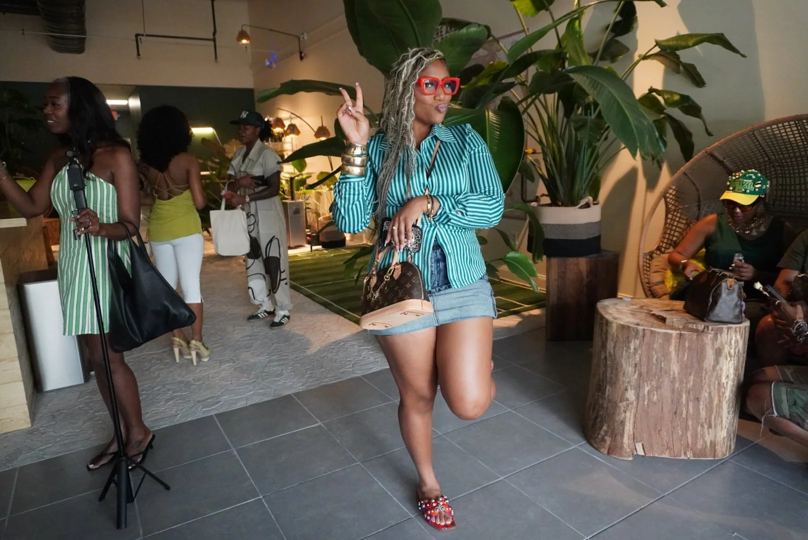 Woman with long braided platinum blonde hair wearing large red glasses, a teal and white striped shirt, denim shorts, and decorative red sandals, posing playfully in a social setting with large green plants and wooden decor.