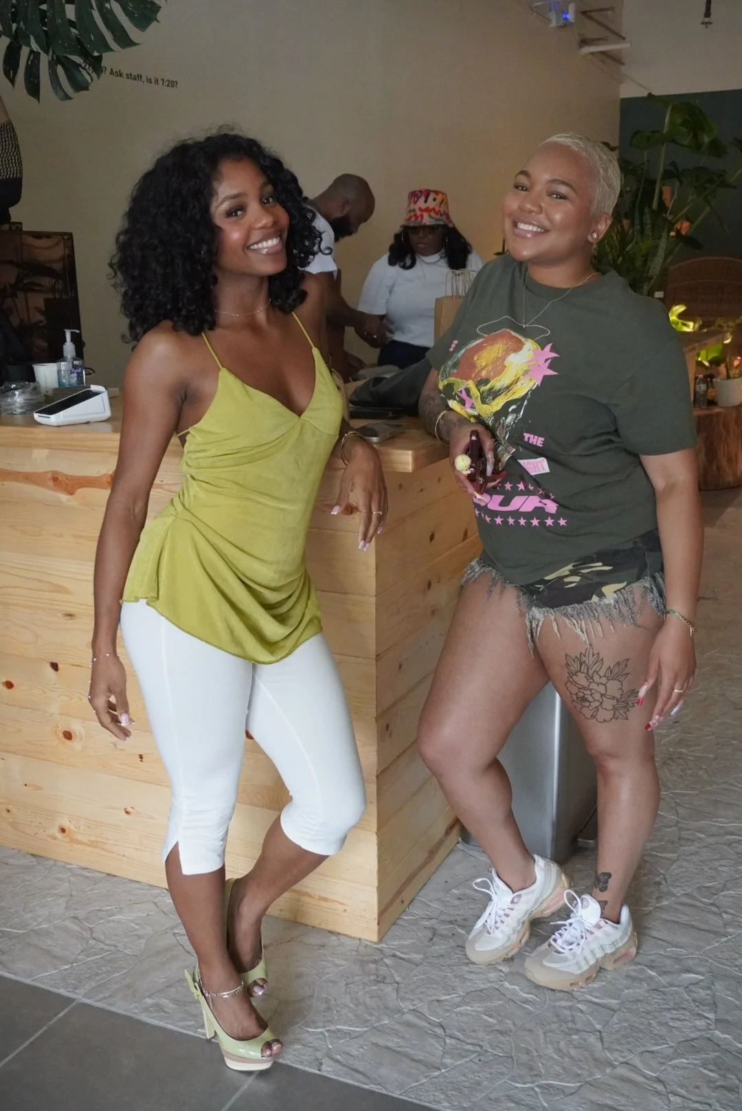 Two women smiling and posing indoors at a wooden counter. One is wearing a yellow top and white pants, the other a graphic t-shirt, camo shorts, and sneakers. Two people are in the background.