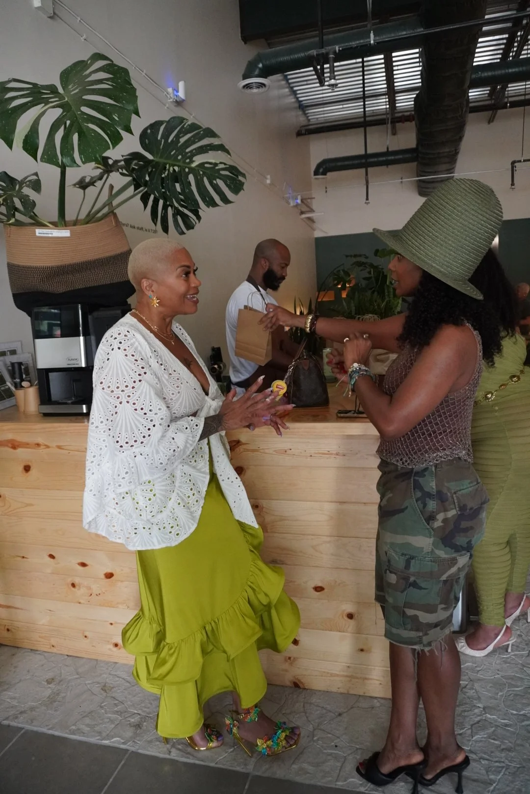 Two women are engaging in conversation at a cafe or restaurant counter. One woman has a platinum-blonde buzz cut, wearing a white eyelet blouse and a lime green skirt, with colorful floral high heels. The other woman has curly hair, wearing a large green sunhat, a fishnet top, and camouflage shorts. A man in the background is holding a paper bag, at the counter with plants and equipment visible around.