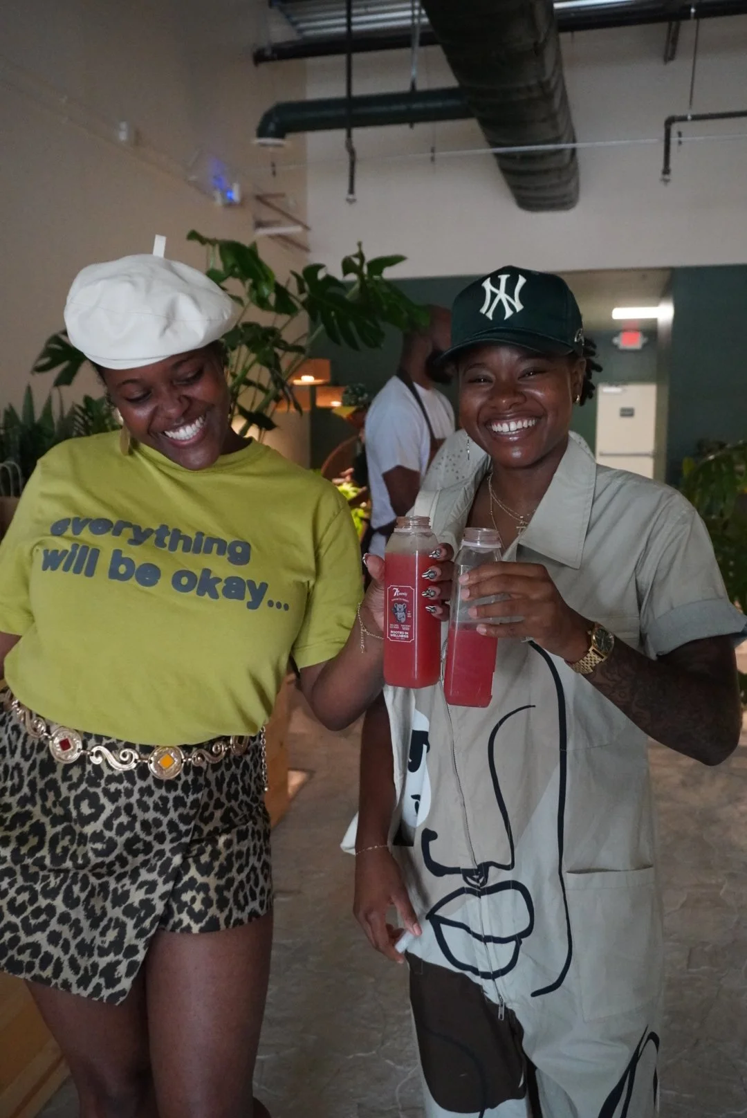 Two women smiling and holding drinks in a social setting. One woman is wearing a white chef's hat and a yellow T-shirt with the message "everything will be okay...". The other woman is wearing a black New York Yankees baseball cap and a beige shirt w