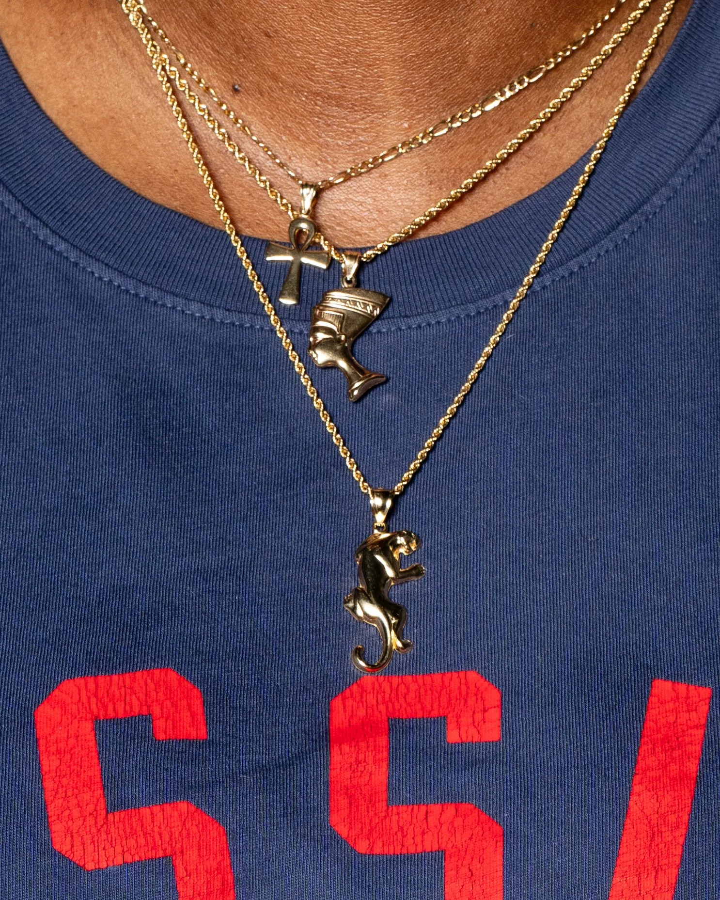 Close-up of a person's neck and chest wearing multiple gold layered necklaces, including an ankh, a head profile, and a Panther, against a blue shirt with red lettering.