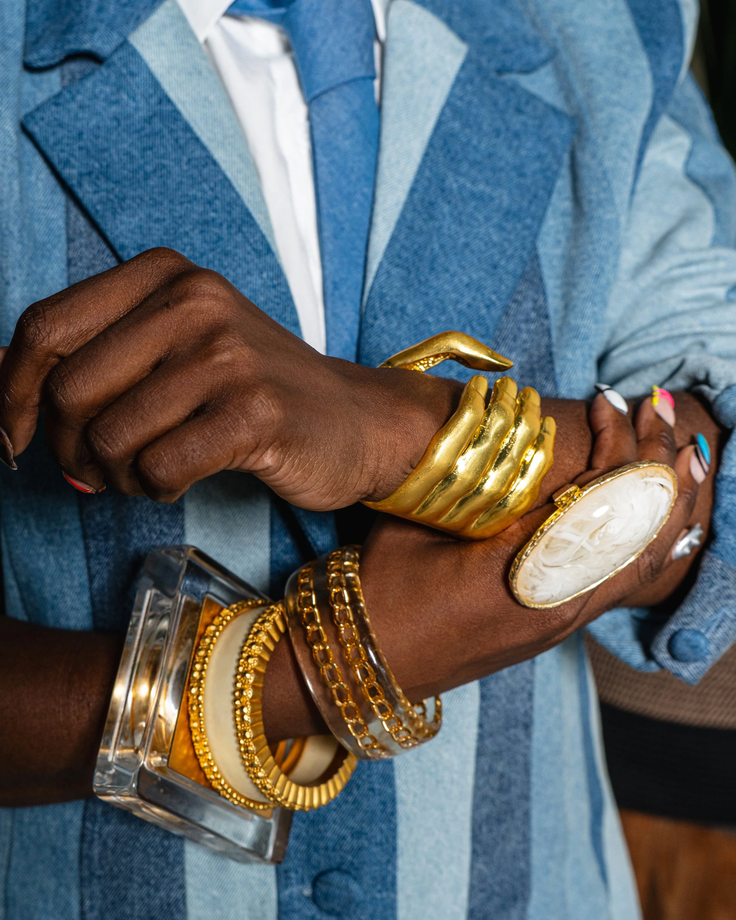Person wearing a denim blazer and white shirt, adorned with multiple gold and silver bracelets and large jewelry accessories on their wrist and hand.