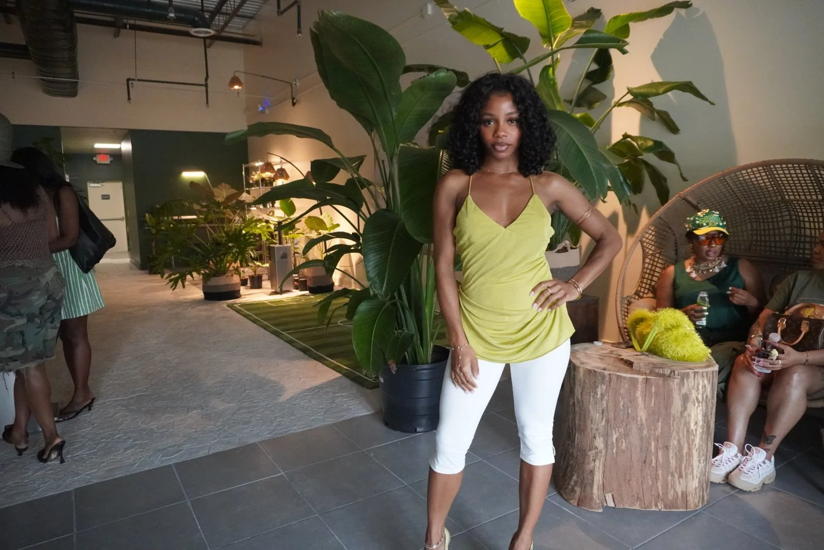 A woman with curly black hair wearing a yellow top and white pants standing next to a large green plant in an indoor lounge area. There are other people sitting and standing in the background.