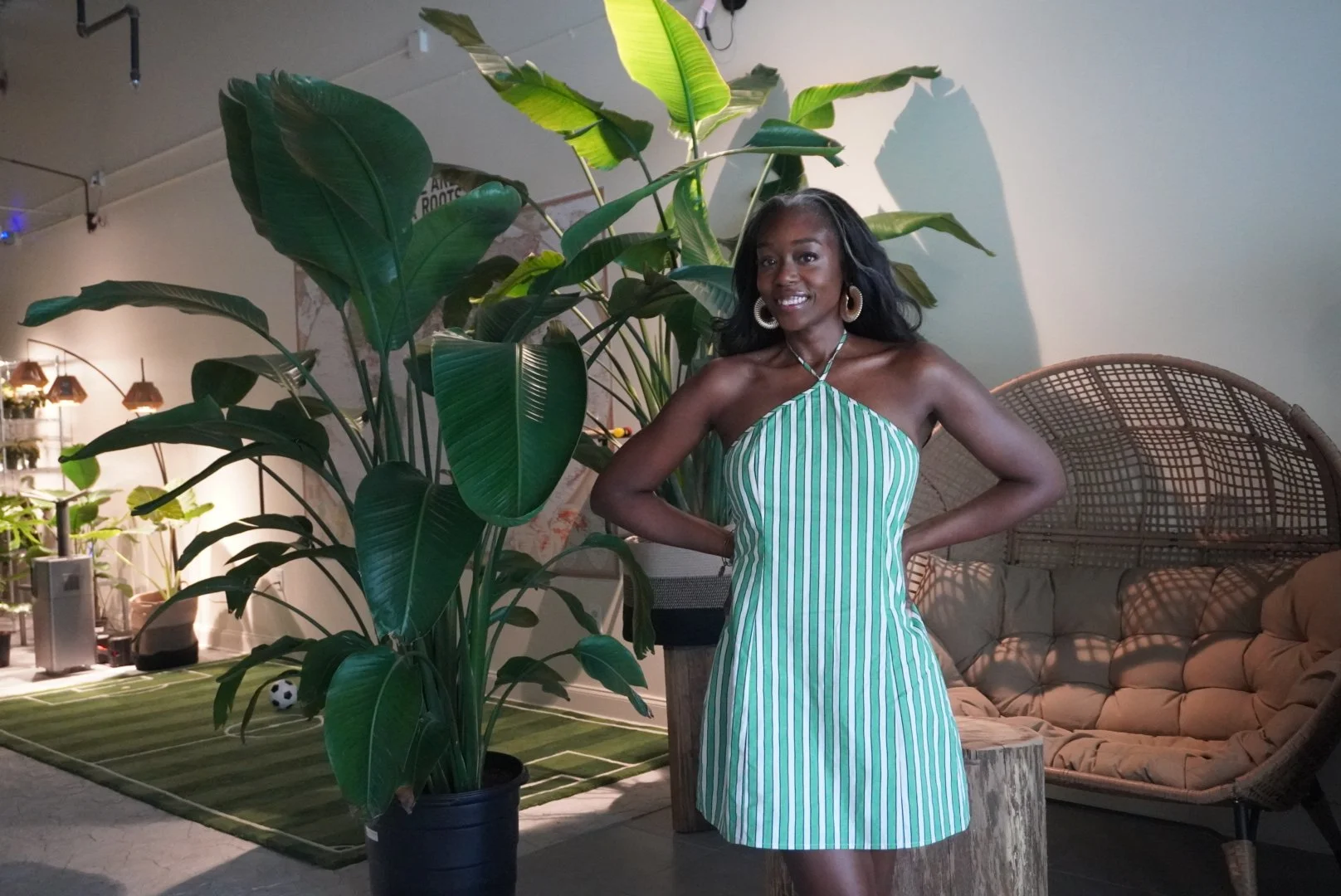 A woman with dark skin and black, shoulder-length hair, smiling and wearing hoop earrings and a green and white striped halter dress, standing indoors next to a large green plant with big leaves. In the background, there is a wicker semi-circular out