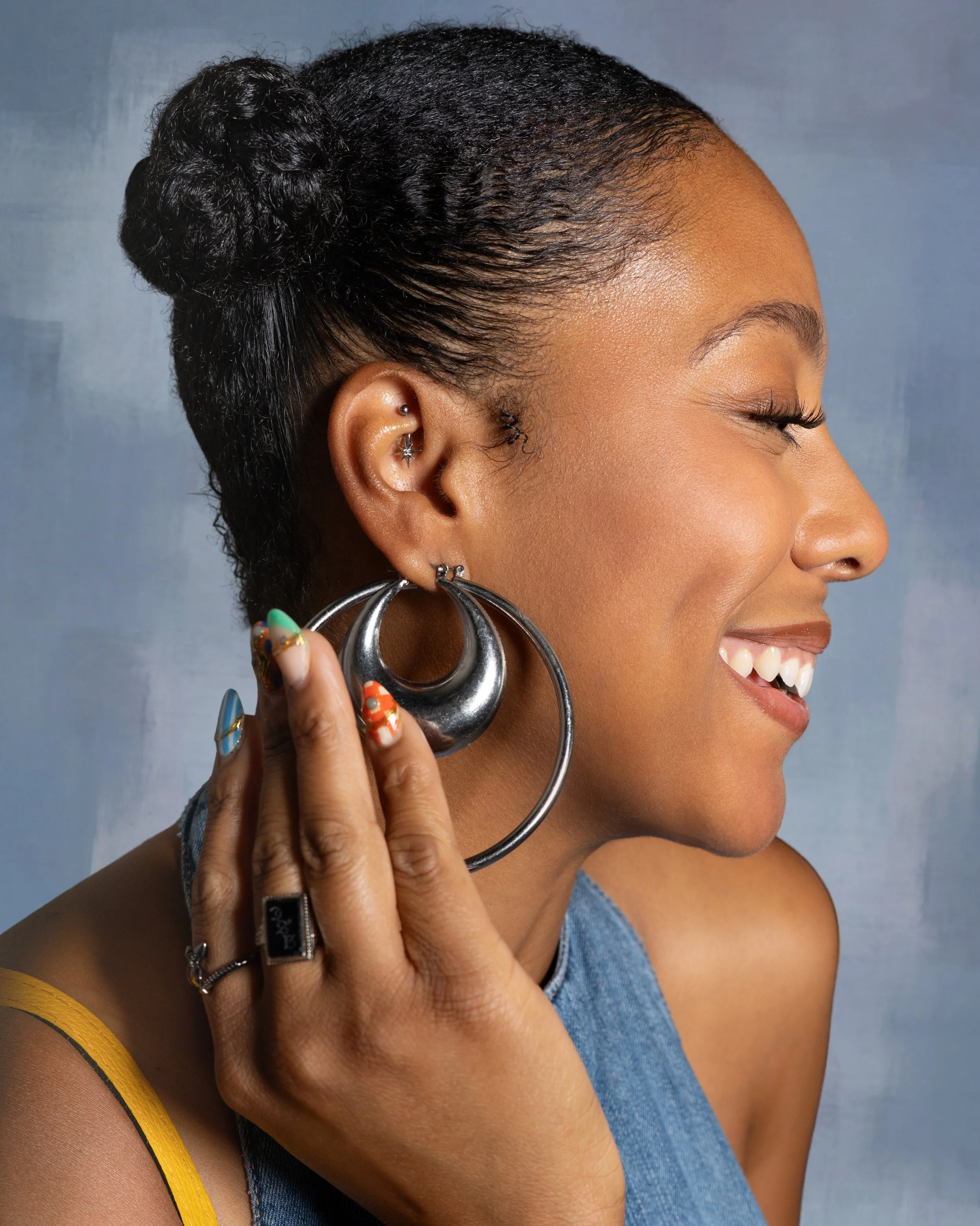 Close-up of a woman smiling with her eyes closed, wearing multiple earrings, holding her ear with her fingers, with a blue background.