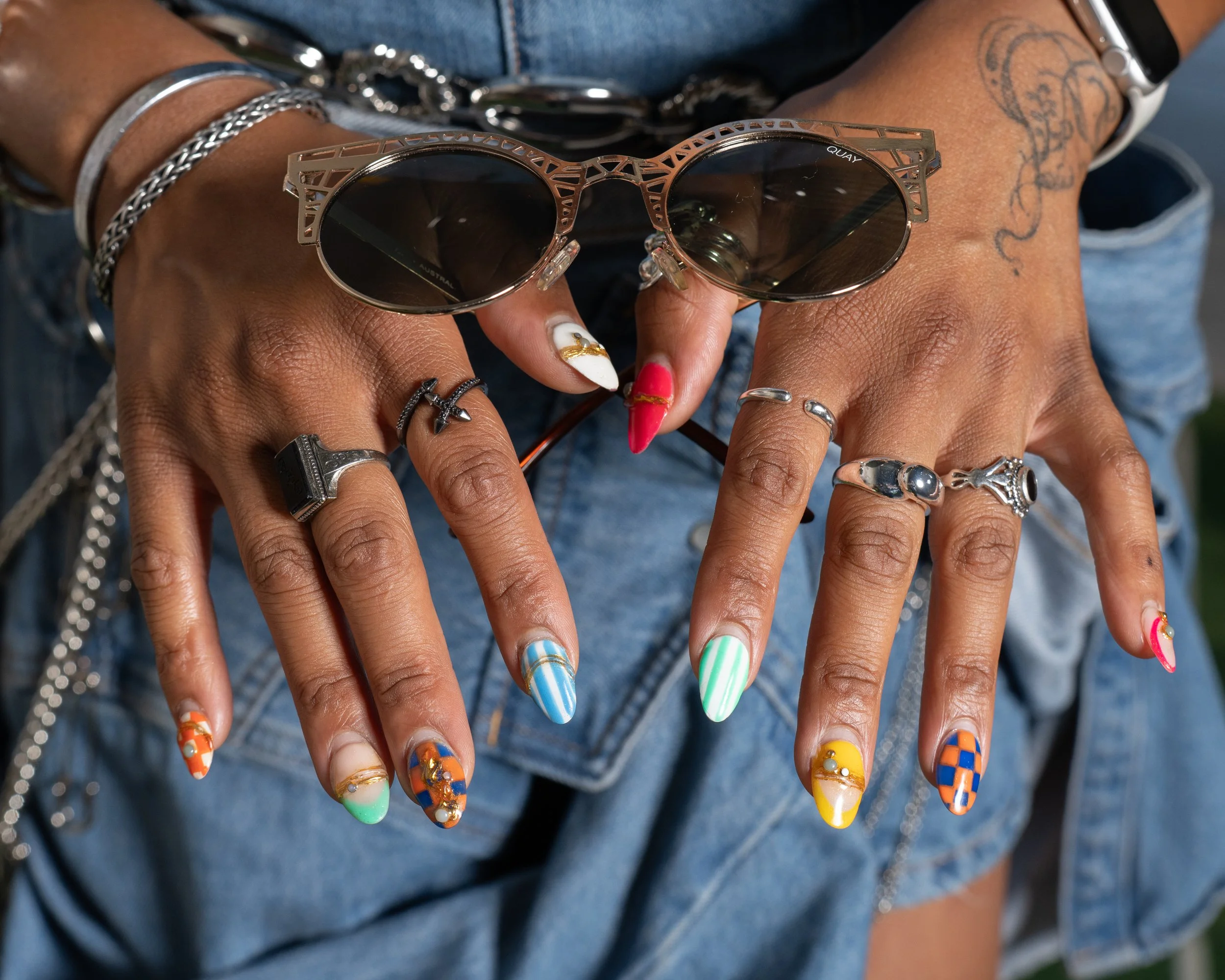 Close-up of a person's hands displaying colorful, patterned long nail art with various nail designs and jewelry, including bracelets, rings, and a tattoo on the arm.