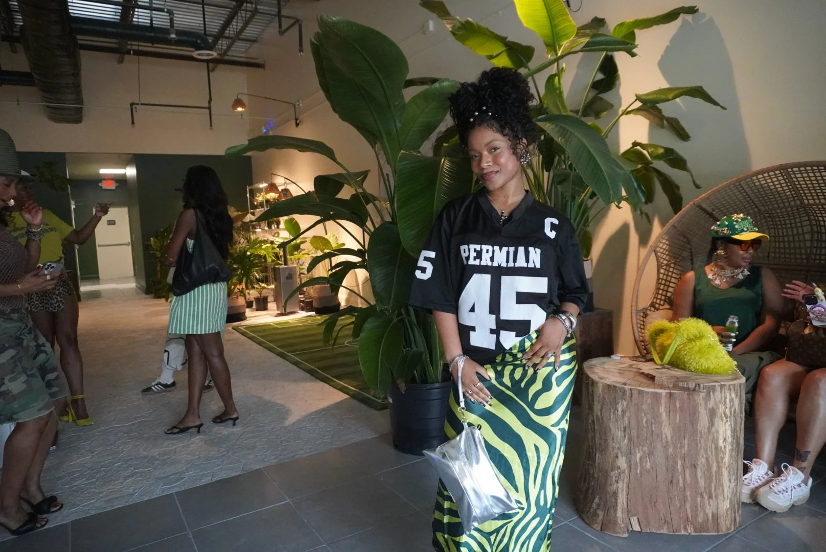 A woman stands in front of large green plants at a social gathering or party, wearing a black sports jersey with white lettering and numbers, and a yellow and black zebra-print skirt.