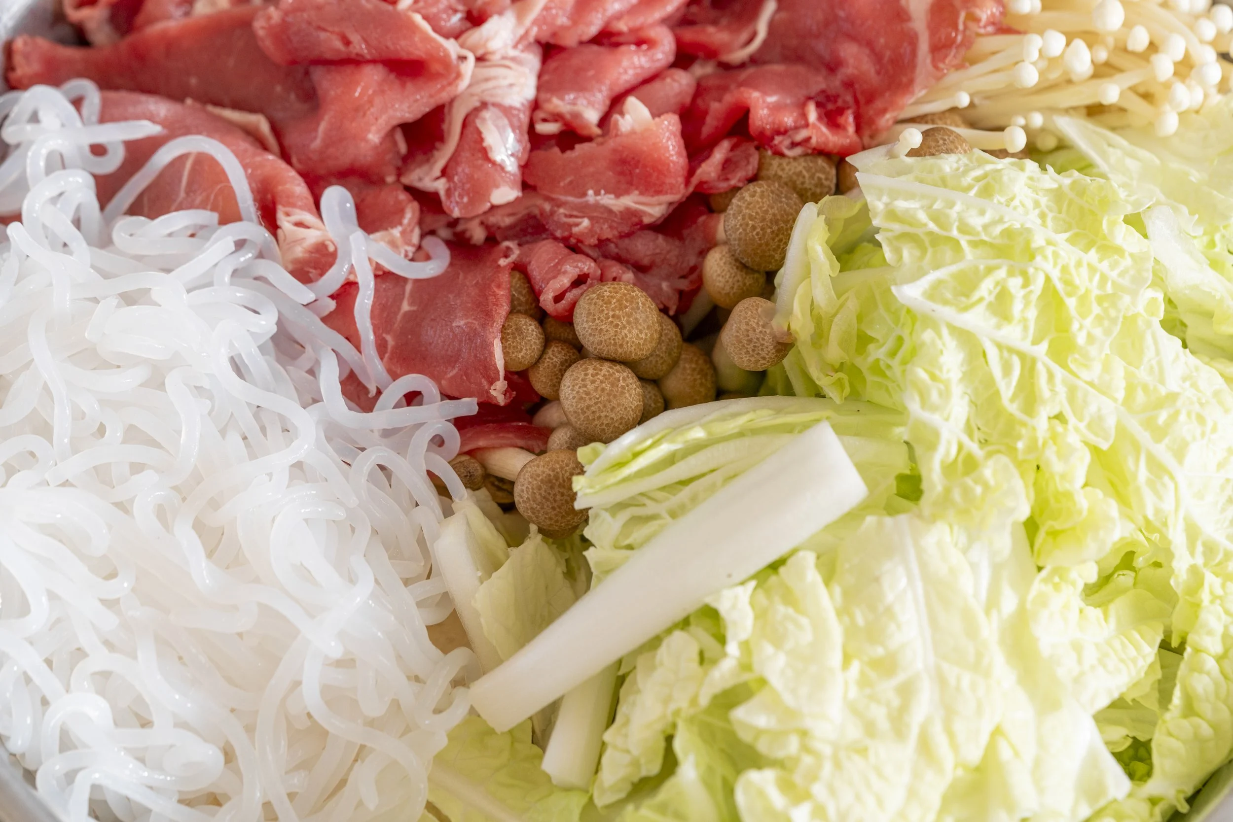 Raw meat, enoki mushrooms, glass noodles, and napa cabbage on a surface, ready for cooking.