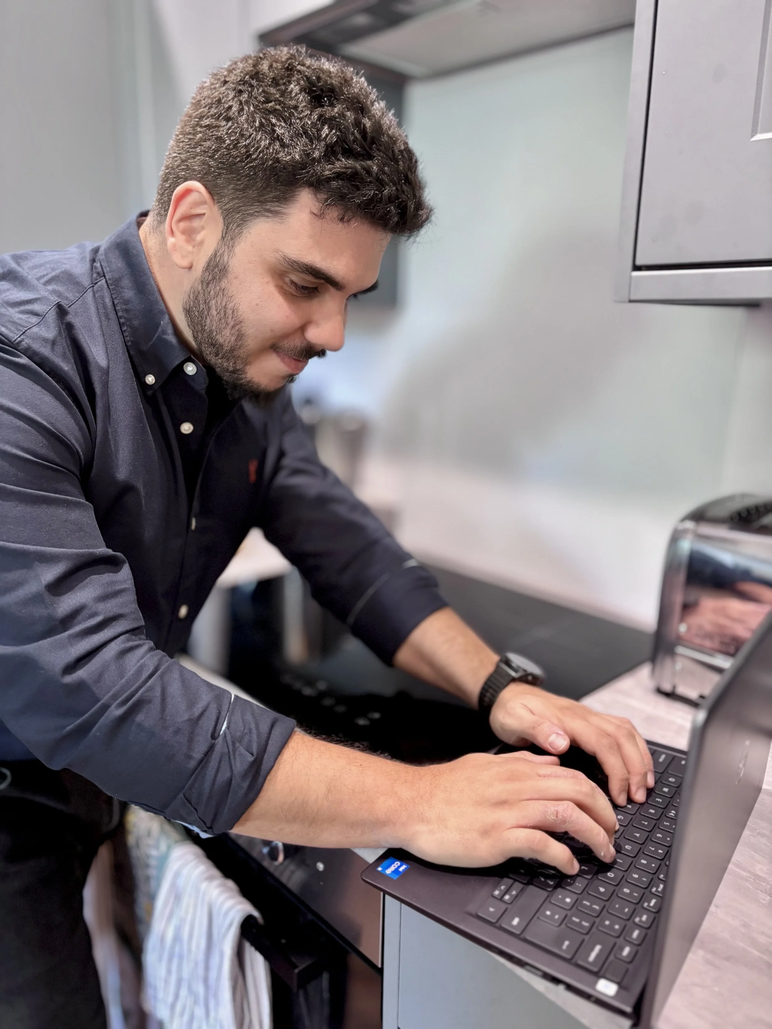 A man with short brown hair and a beard working on a laptop at a desk in an office or classroom setting.