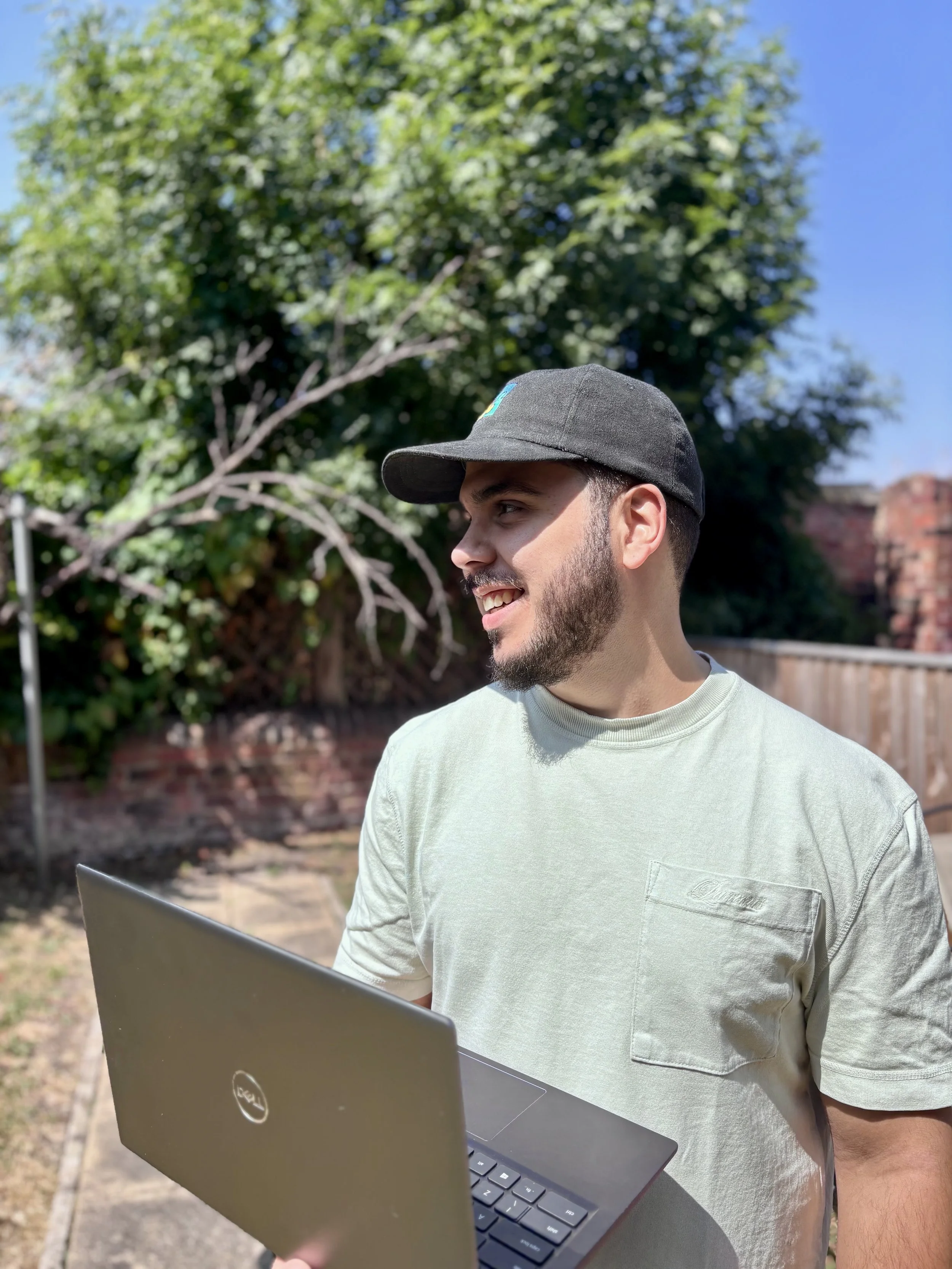 Young man with a beard in a gray t-shirt and a gray baseball cap using a laptop outdoors, with trees and a brick wall in the background on a sunny day.