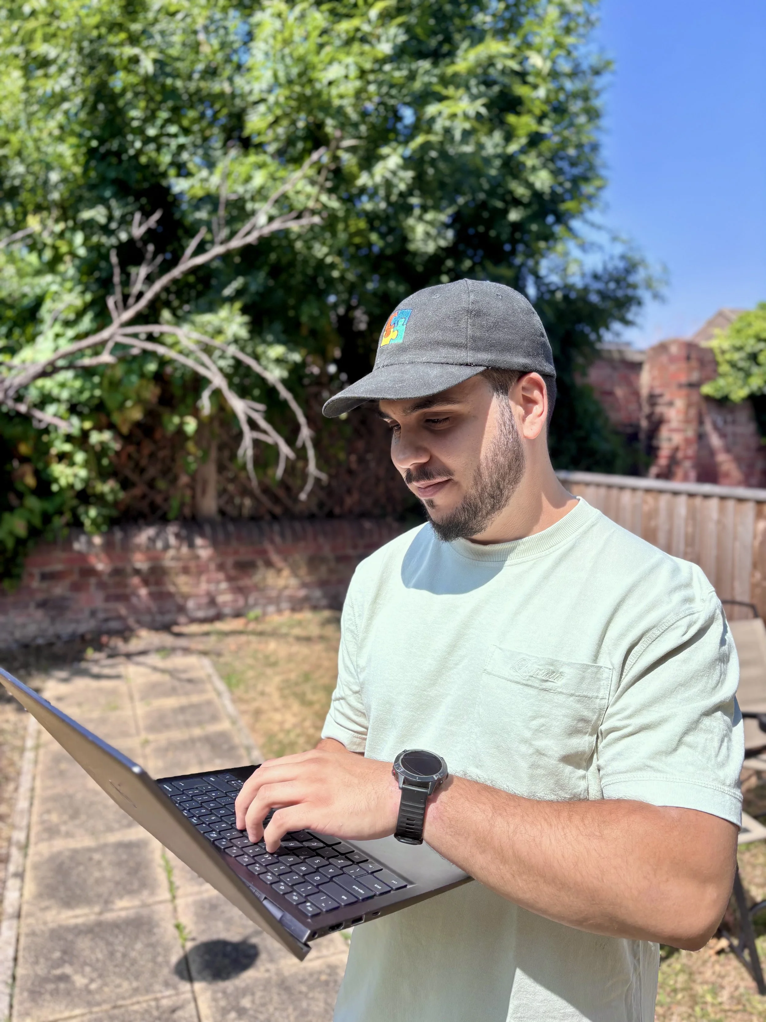 A young man outdoors, wearing a gray cap with a colorful puzzle piece logo, a light green t-shirt, and a smartwatch, is typing on a laptop. Trees and a brick wall are in the background under a clear blue sky.