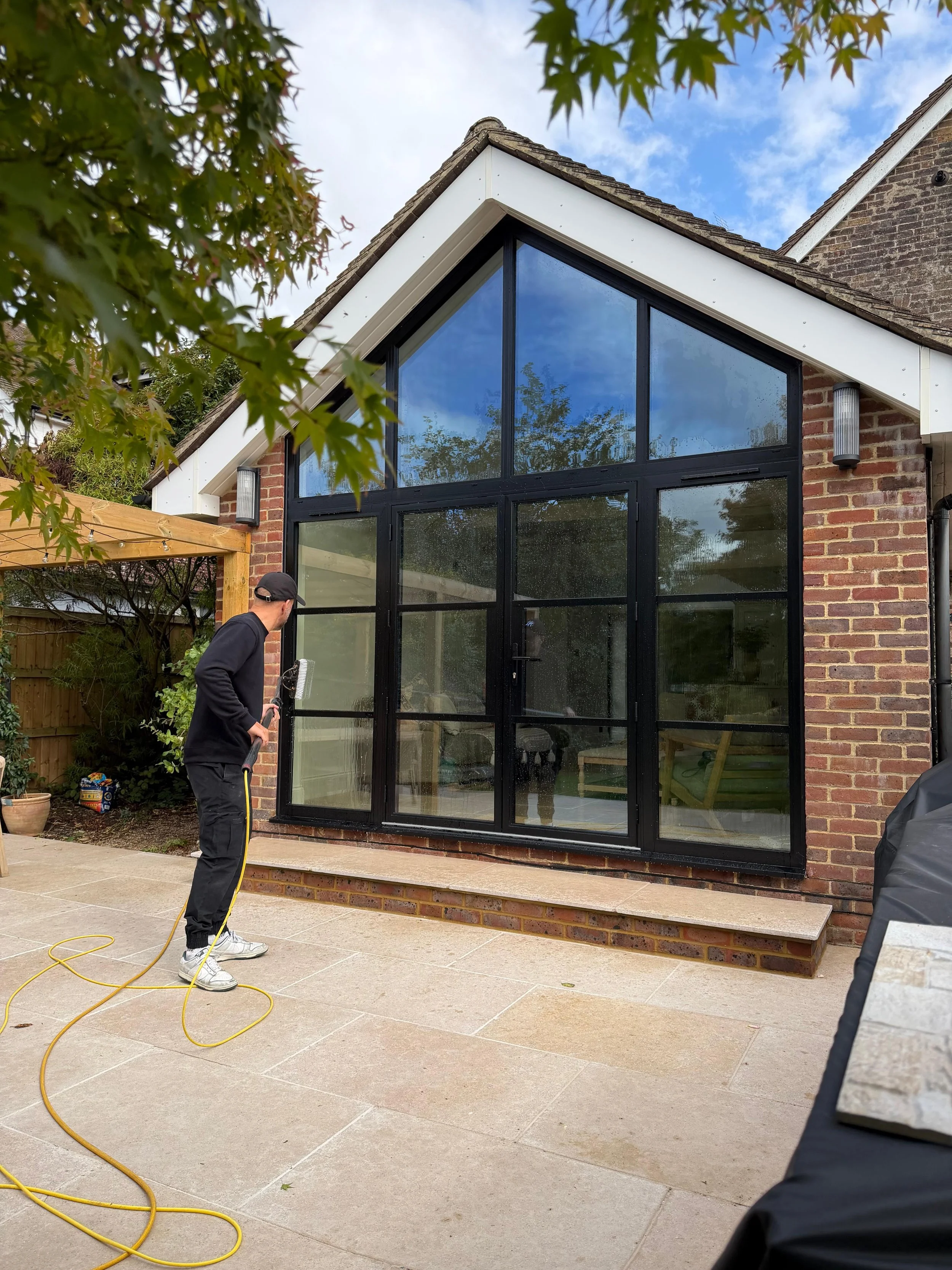 A person cleaning exterior glass doors of a modern house with a spray gun, in a backyard with trees, a wooden trellis, and a brick wall.
