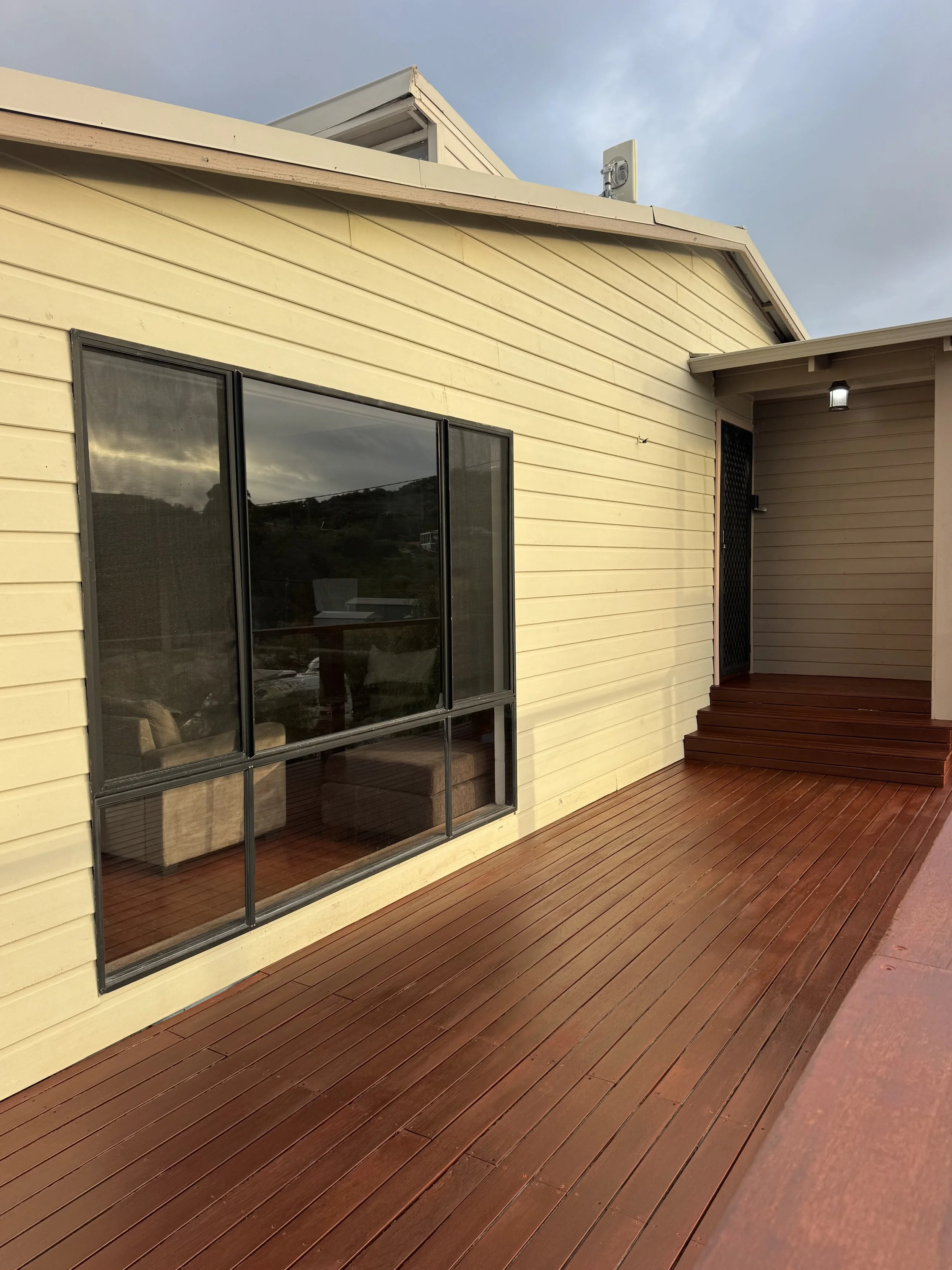 A house with beige wood siding and dark window frames, featuring a sliding glass door leading to a wooden deck, with a small covered area and a light fixture, under a cloudy sky.