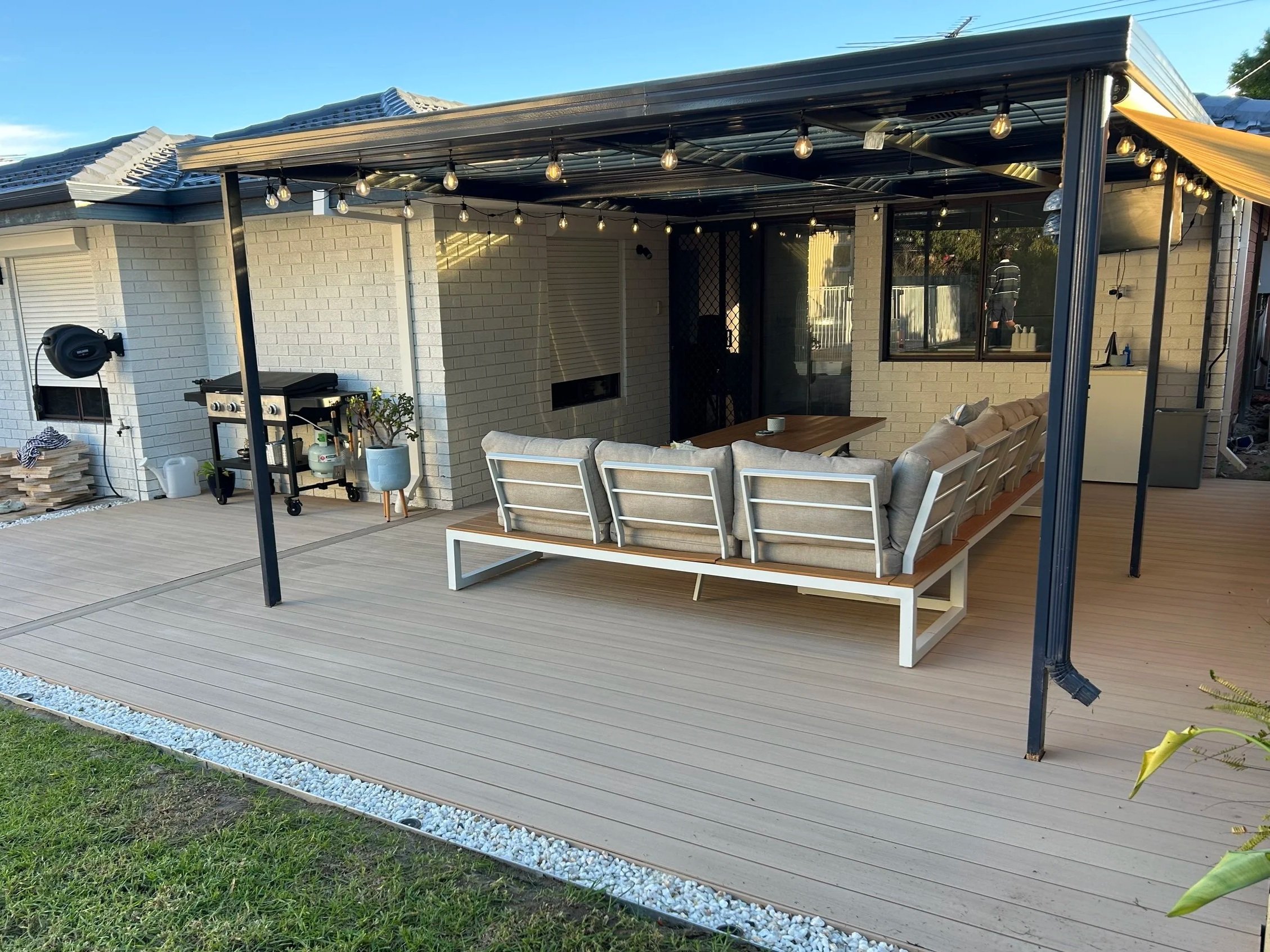 An outdoor patio area with beige cushioned chairs, a wooden table, string lights, a grill, and a house with brick walls and sliding glass doors.