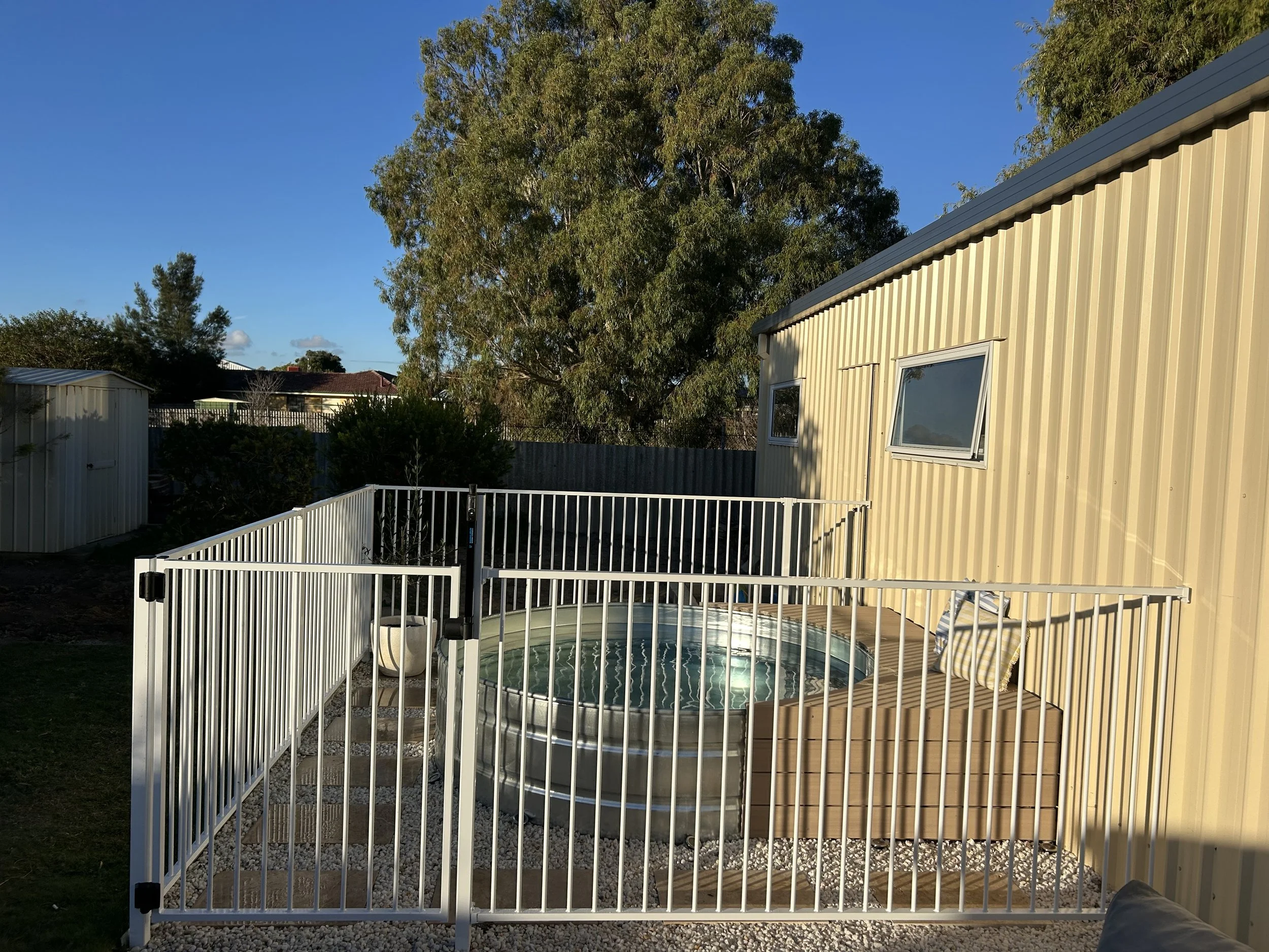 A backyard with a cream metal shed, a round above-ground pool with a white fence, a lounge chair with striped cushions, tall trees, and neighboring houses under a clear blue sky.