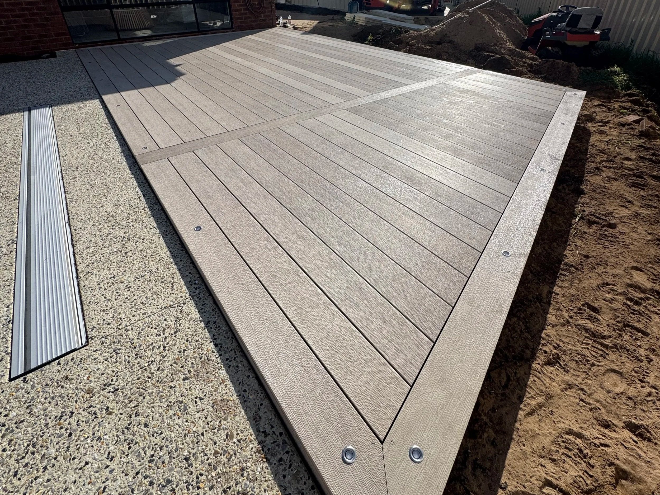 View of a wooden deck next to a sliding glass door, with construction area and dirt on one side, and a used tool box in the background.