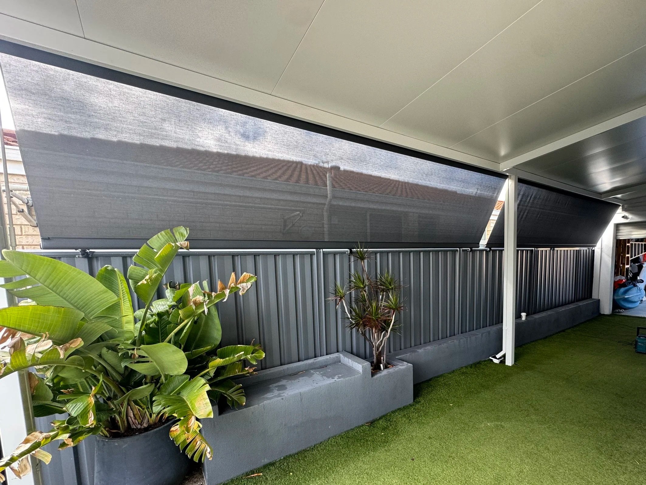 A backyard patio with a gray metal fence, potted plants, and a shade sail over the area. Green grass or artificial turf covers the ground.