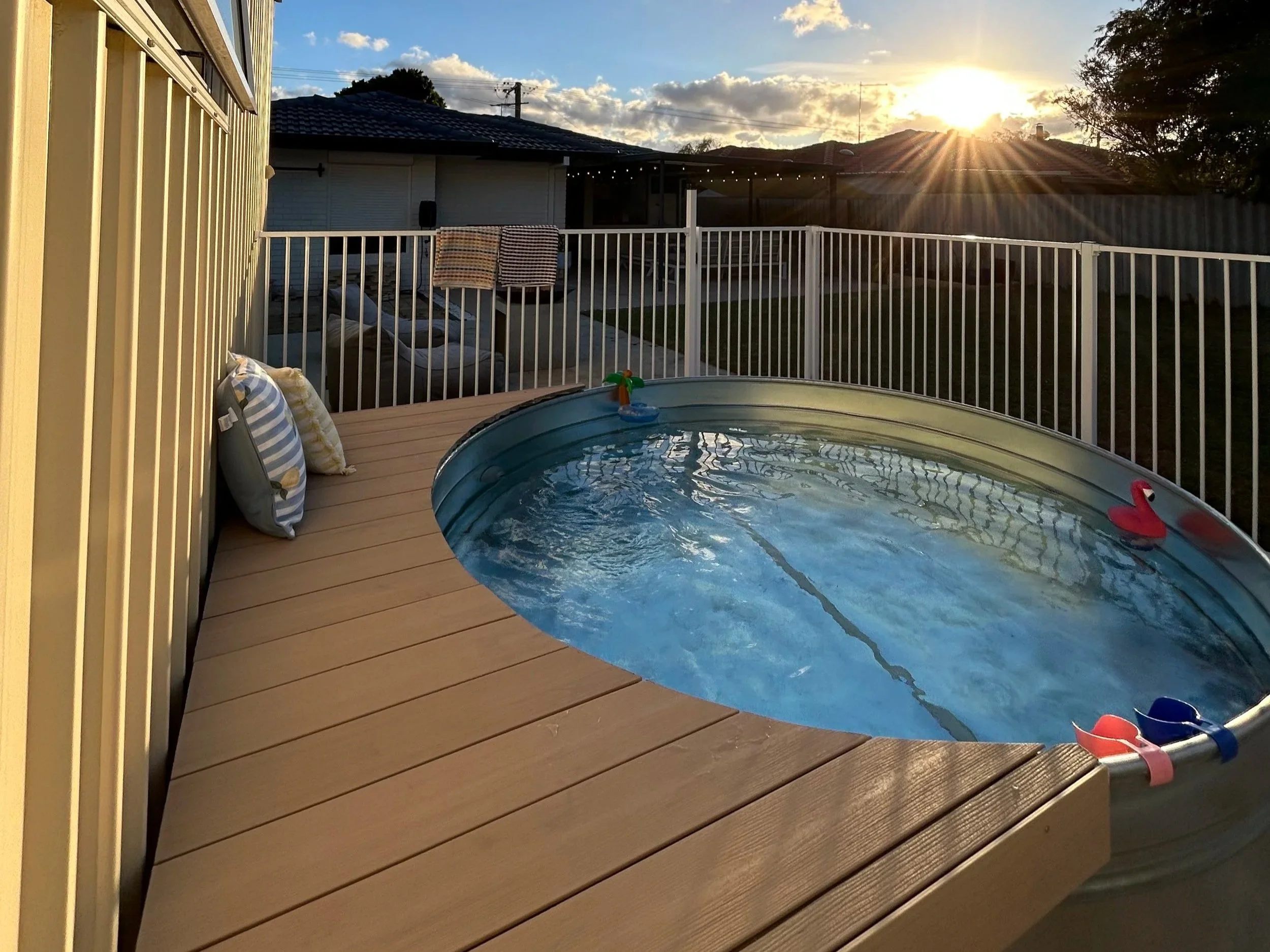 A backyard deck with a small round pool, a lounge chair with pillows, and clothes hanging on a railing. The sun is setting, casting a warm glow and sun rays across the sky, with houses, trees, and utility poles in the background.