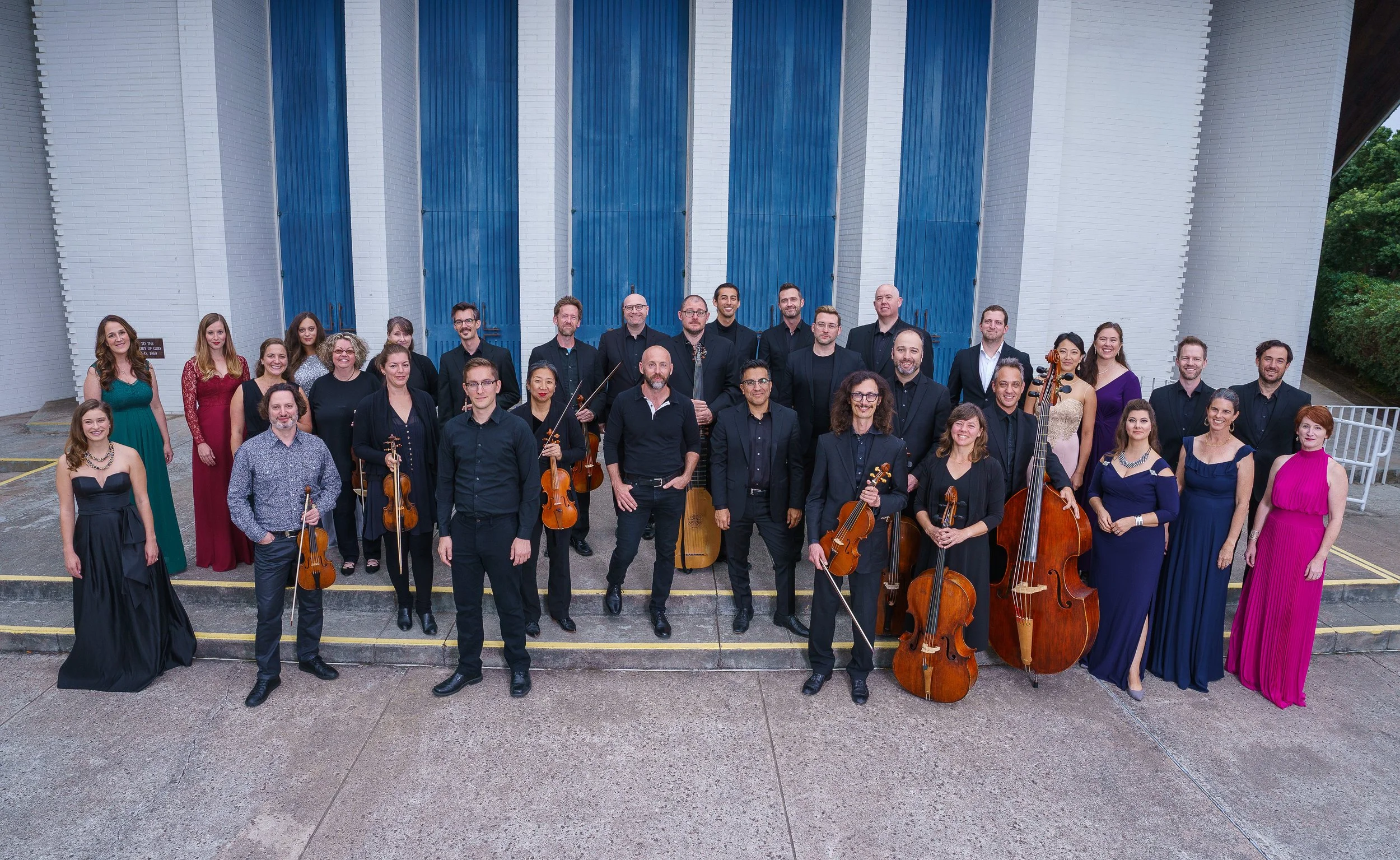 Group of musicians and singers posing outside a building with blue doors, holding string instruments, and dressed in formal attire.
