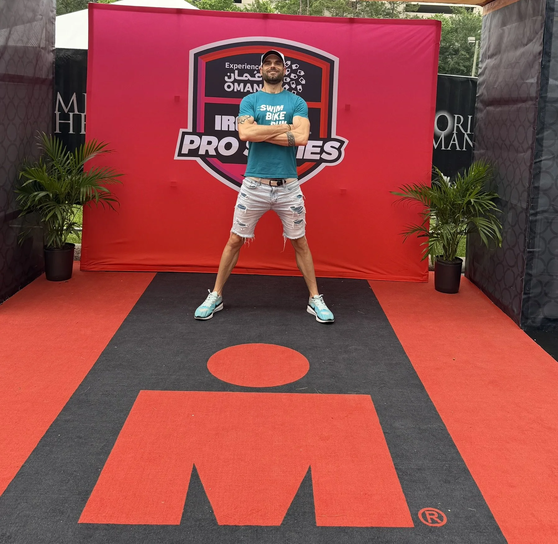 A man with a beard and tattoos wearing a turquoise shirt, ripped shorts, and running shoes stands with arms crossed on a black and red mat in front of a pink backdrop with a logo that reads "Oman" and "Pros" surrounded by green plants.