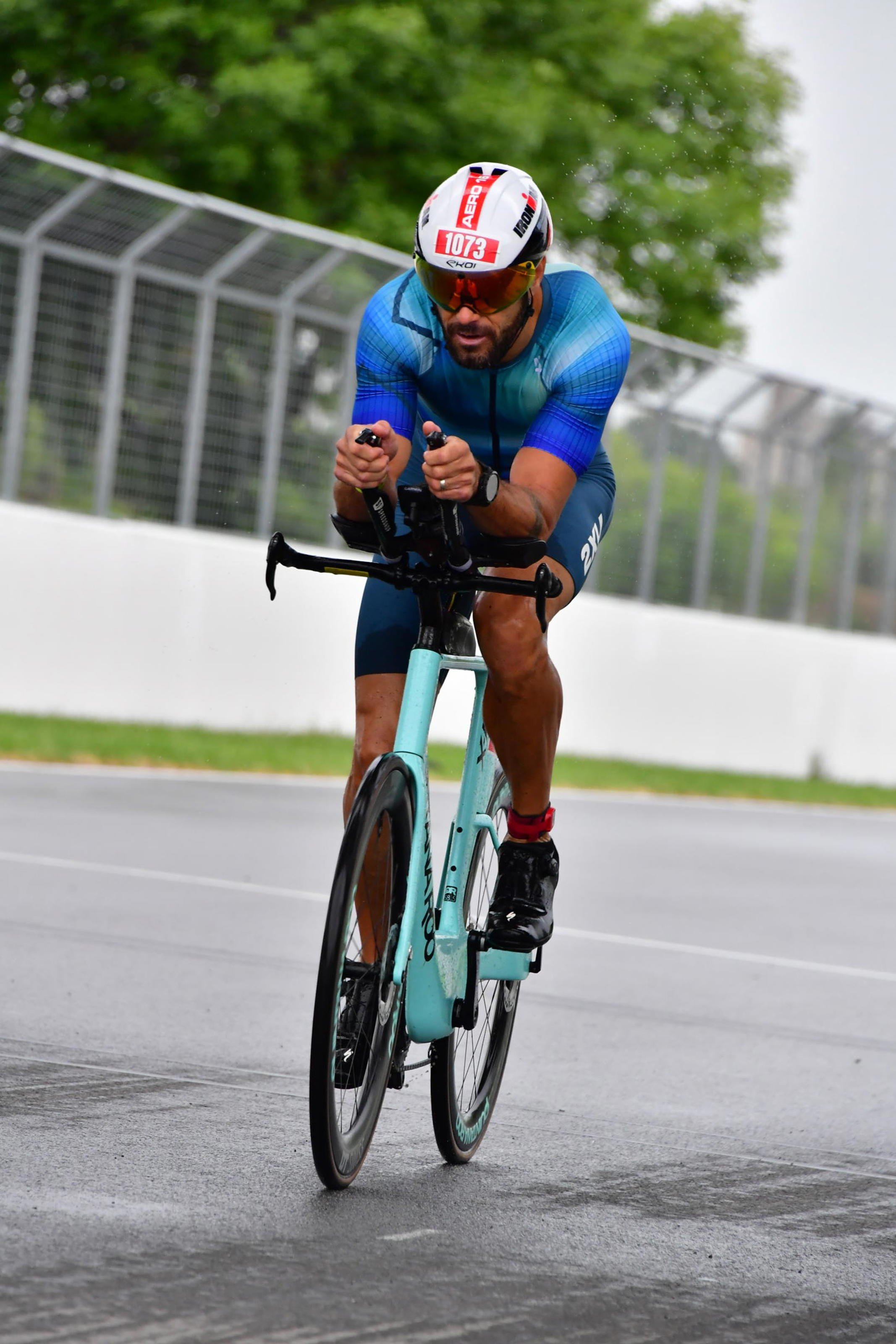 A cyclist wearing a blue jersey, black shoes, and a white helmet with the number 1073 on it, riding a light blue bicycle on a road bordered by a fence and trees.