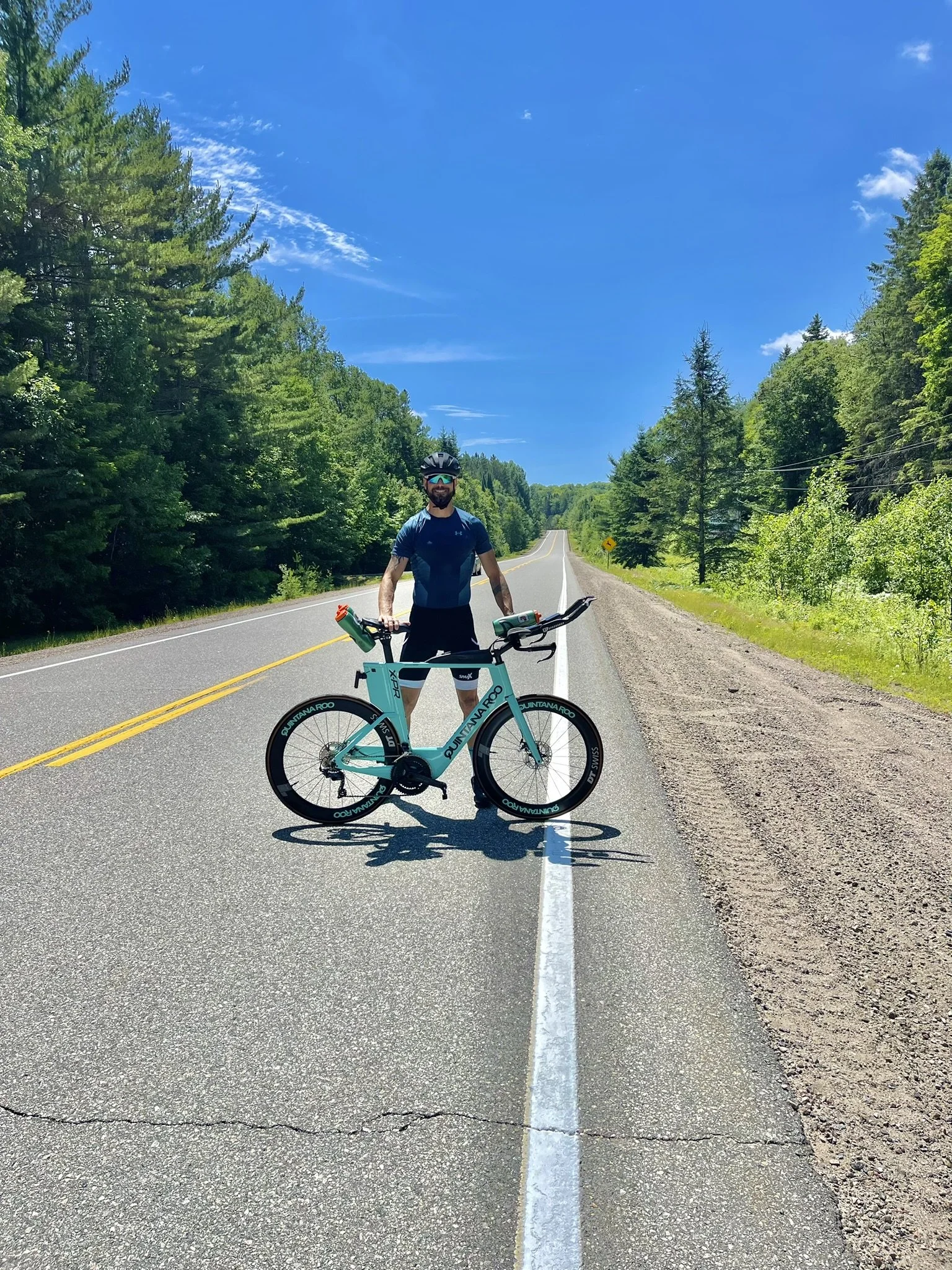 A man wearing a helmet and sunglasses stands with a blue bicycle on a two-lane road surrounded by dense green trees under a bright blue sky with some wispy clouds.