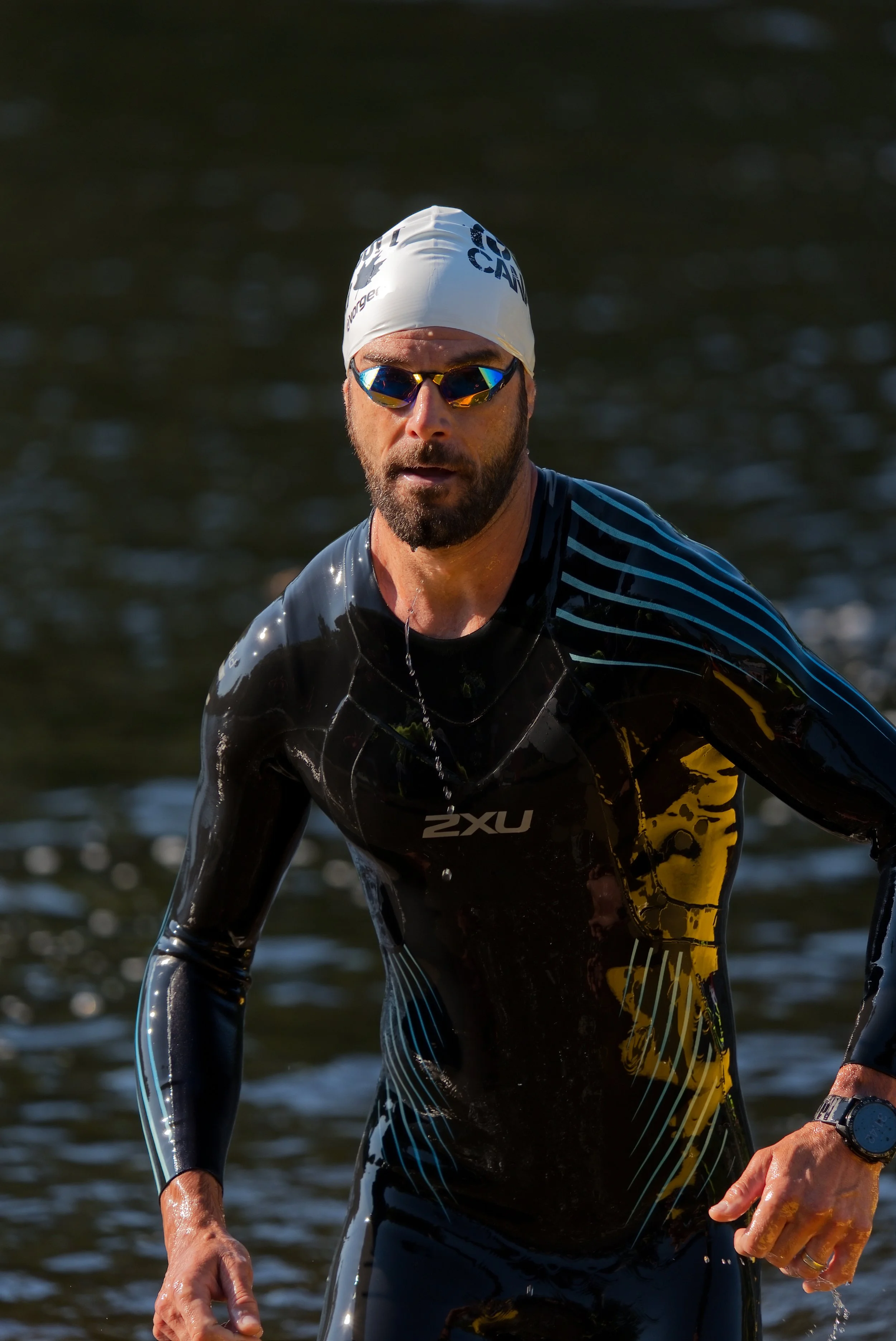 A man wearing a white swim cap, sunglasses, and a black and yellow wetsuit, emerging from water.