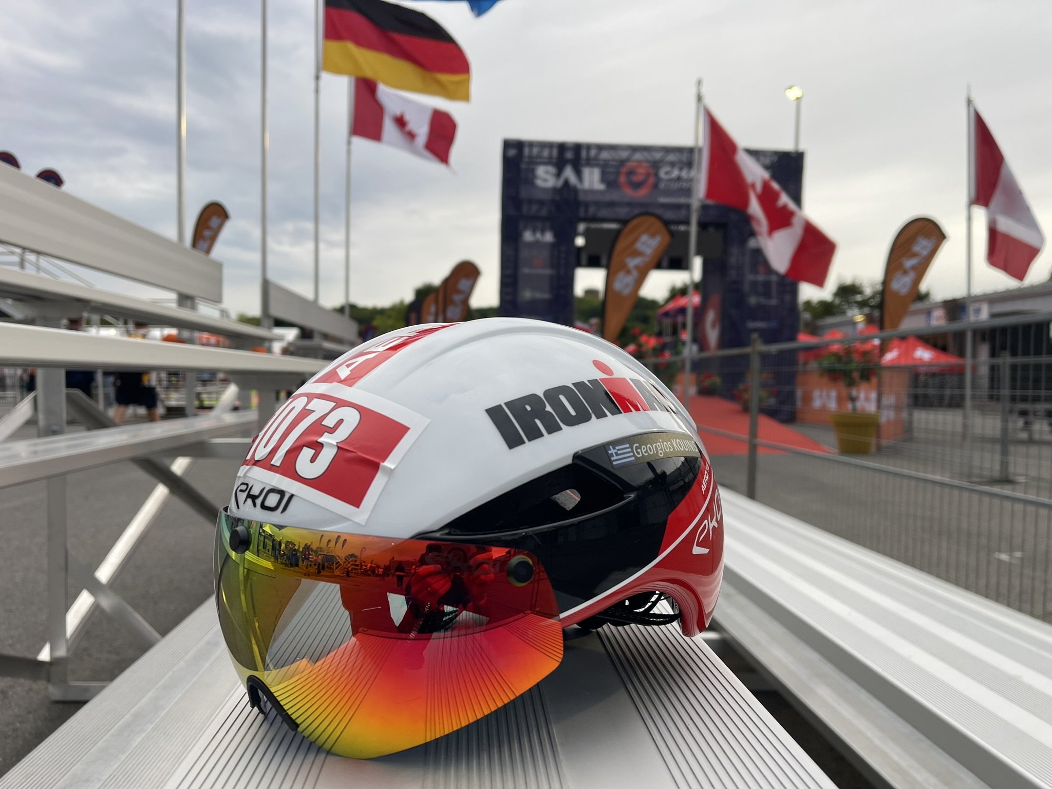 Close-up of a racing helmet with a red, white, and yellow design, placed on a metal surface at a racing event with flags and banners in the background.
