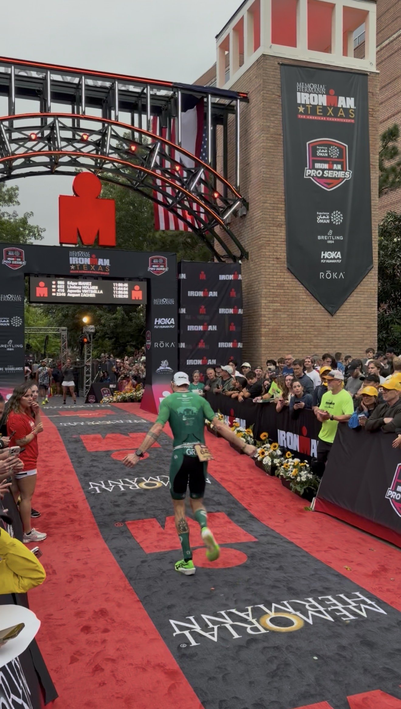 A male athlete wearing a green shirt and black shorts finishing a race at the Ironman Texas competition, crossing the red and black finish line with a crowd of spectators on the sides.