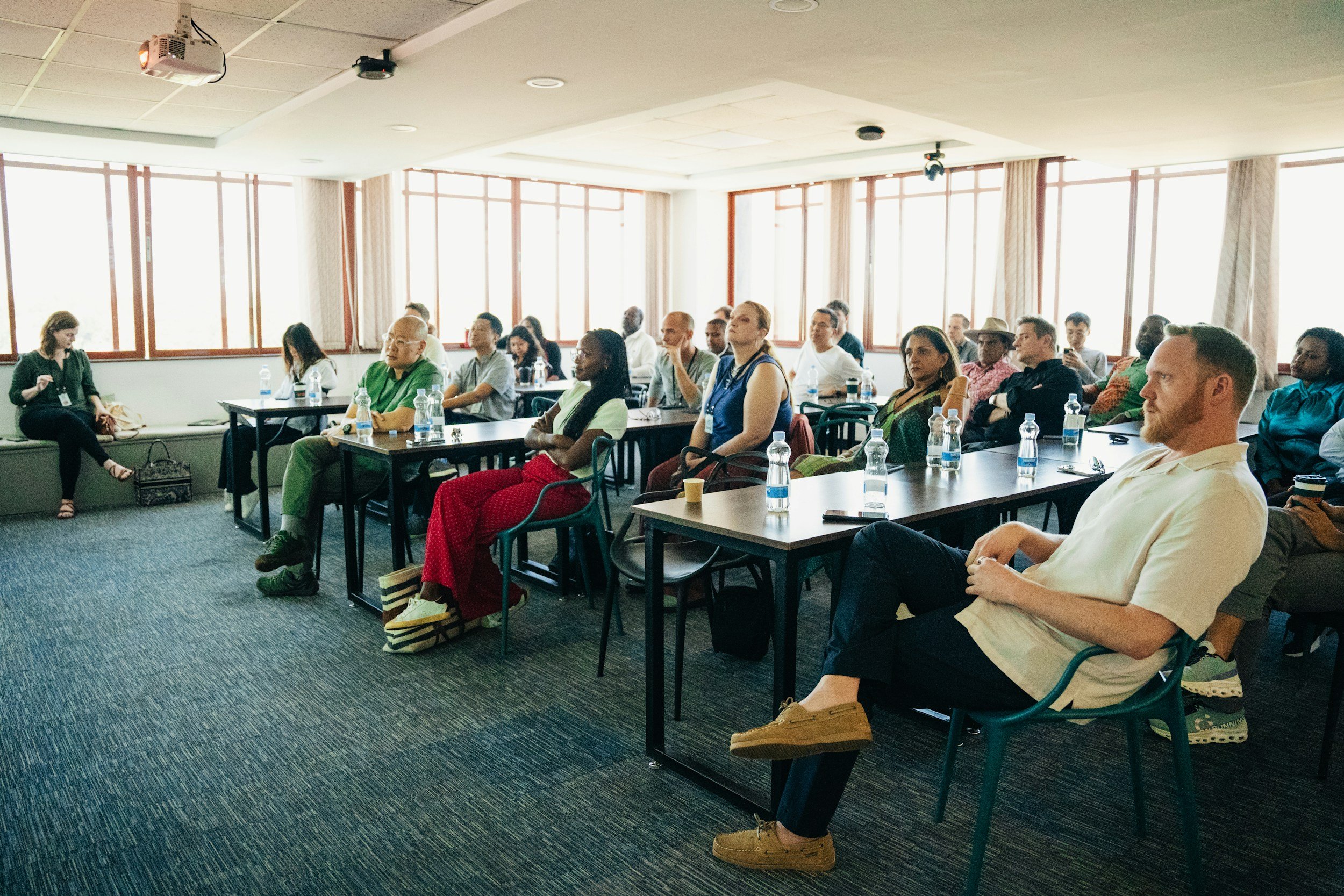 A diverse group of people attending a conference or seminar in a well-lit room with large windows, sitting at tables with bottled water, listening to a speaker or presentation.
