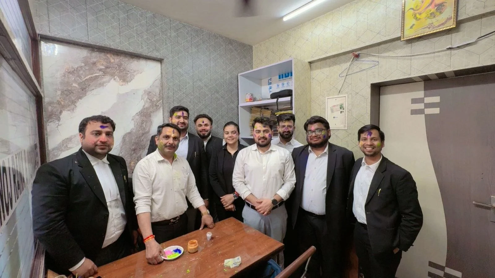Team of nine people dressed in formal attire celebrating with colorful powders on their faces in chamber.