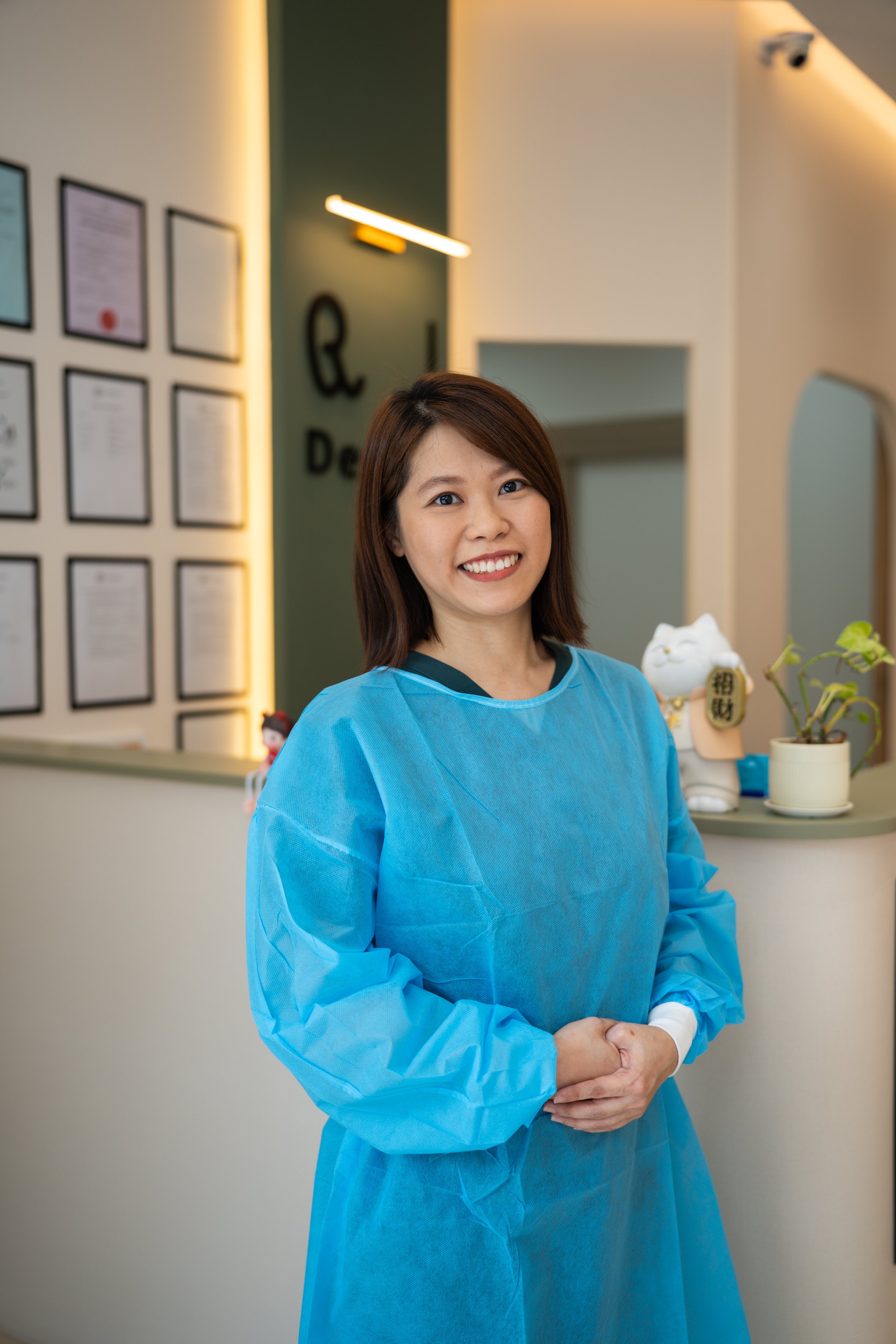 A woman in a blue protective gown smiling with her hands clasped in front of her in an indoor setting, with framed certificates and decor on the wall behind her.