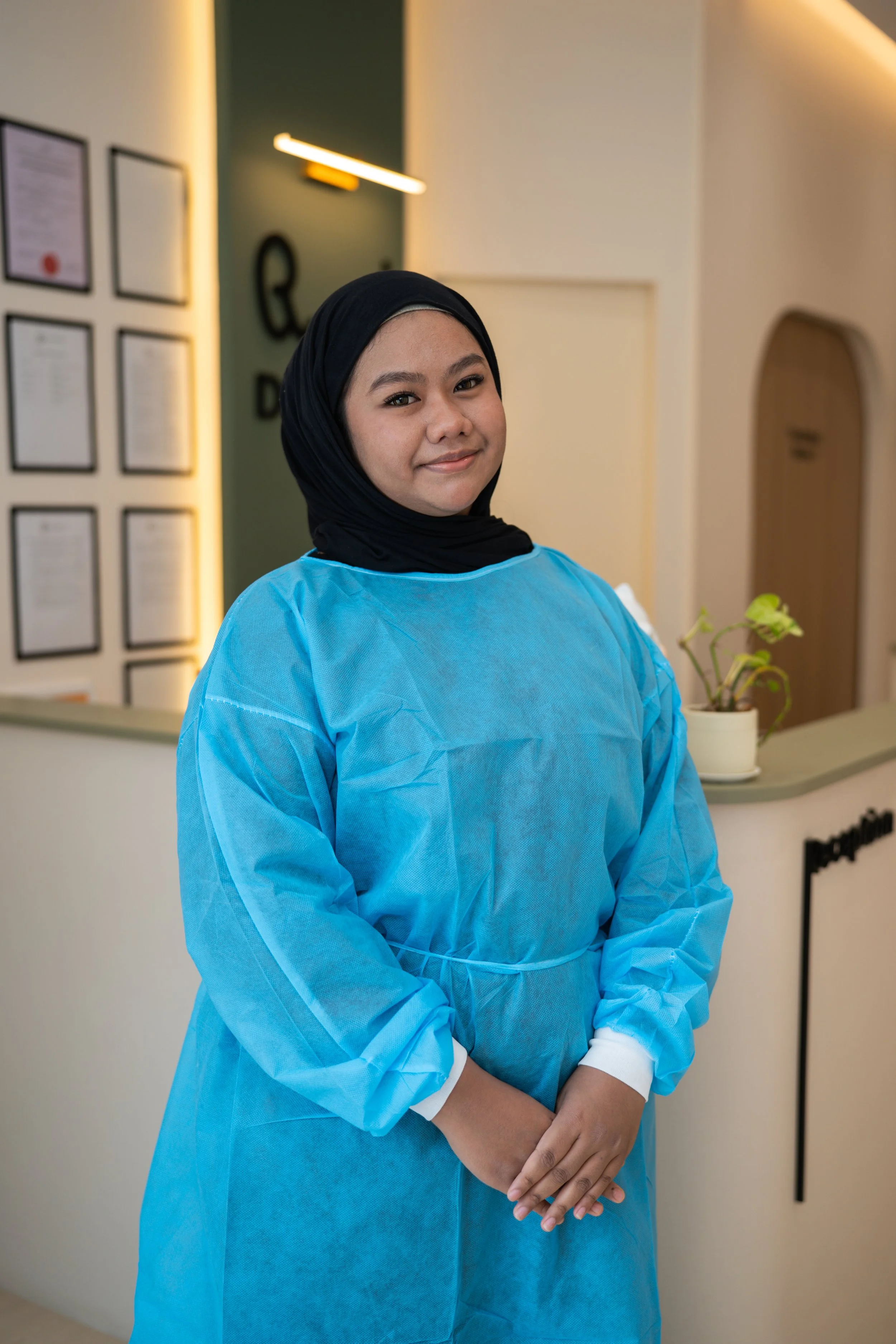 A woman wearing a black hijab, blue medical gown, and white gloves standing in a medical or healthcare facility with framed documents on the wall and a small potted plant in the background.