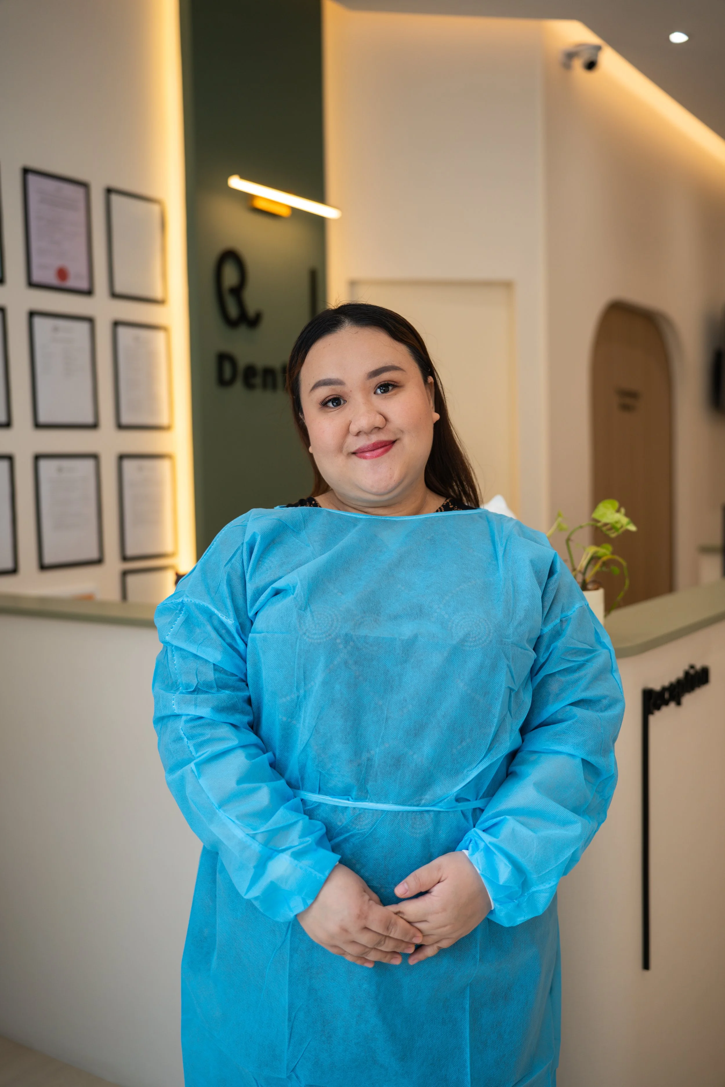 A woman in a blue medical gown standing in a modern clinic or healthcare facility with a friendly expression.