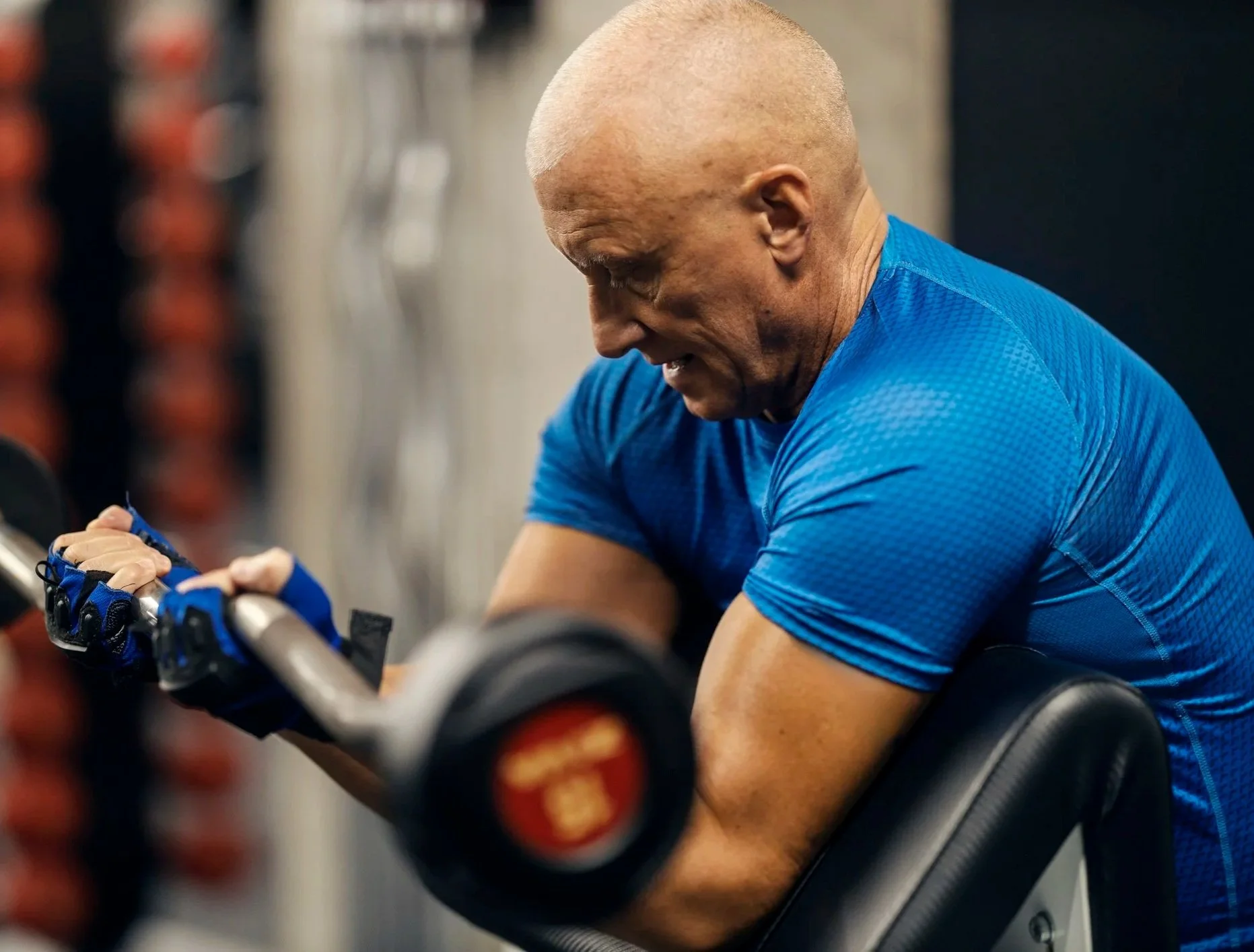 A muscular man with a shaved head in a blue workout shirt lifting weights in a gym.  Personal Training for the over 50's. Ramsbury, Wiltshire.