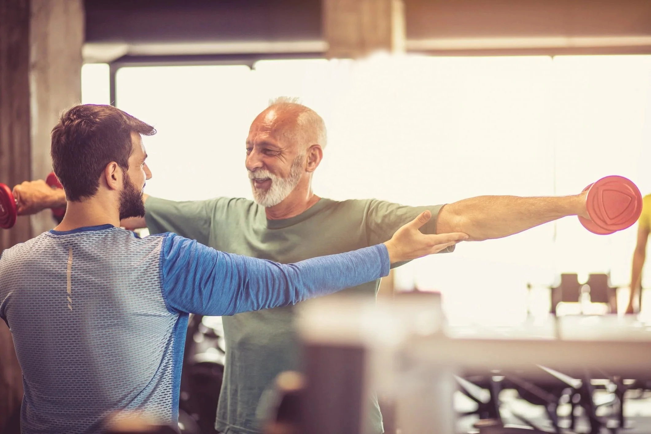 A senior man and a younger man smiling during a workout session at the gym. The senior man is extending his arm with a dumbbell and the younger man is assisting or supporting him.  Personal Training for the over 50's. Ramsbury, Wiltshire.