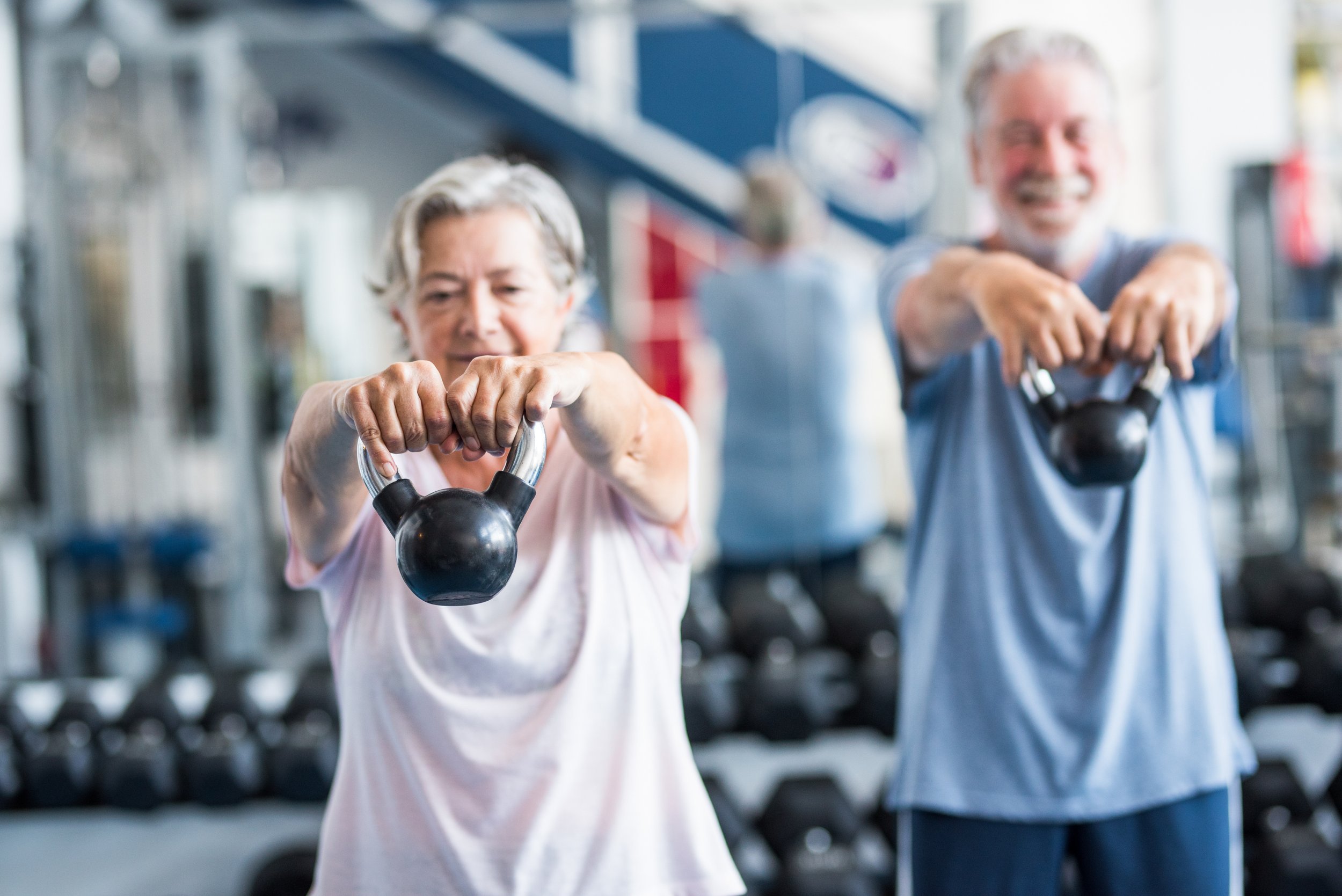 Older woman and older man exercising with kettlebells in a gym, smiling and looking at the camera.  Personal Training for the over 50's. Ramsbury, Wiltshire.