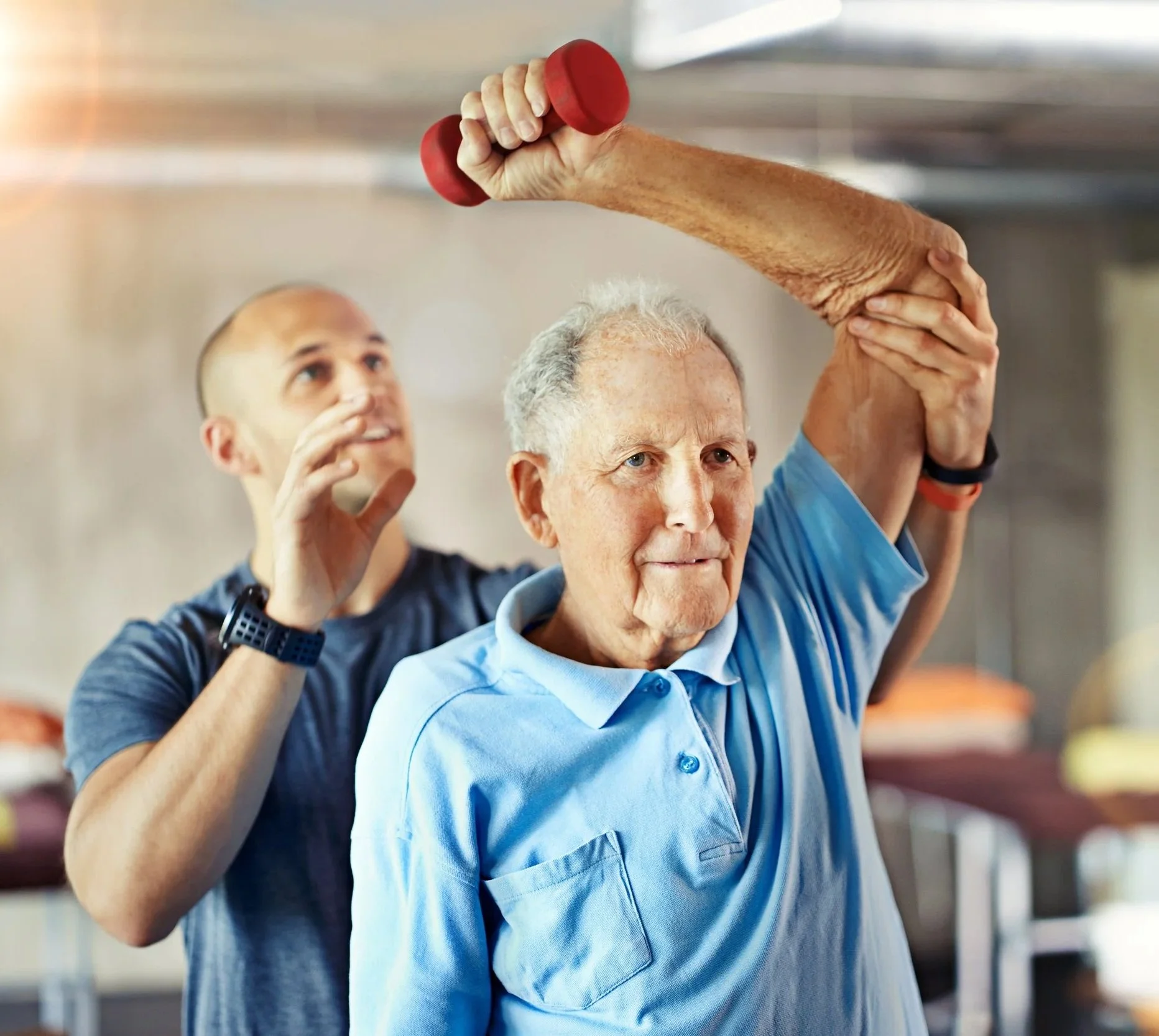 An elderly man with gray hair and a blue shirt participating in a stretching exercise with the assistance of a younger man in a dark shirt, in an indoor setting.  Personal Training for the over 50's. Ramsbury, Wiltshire.