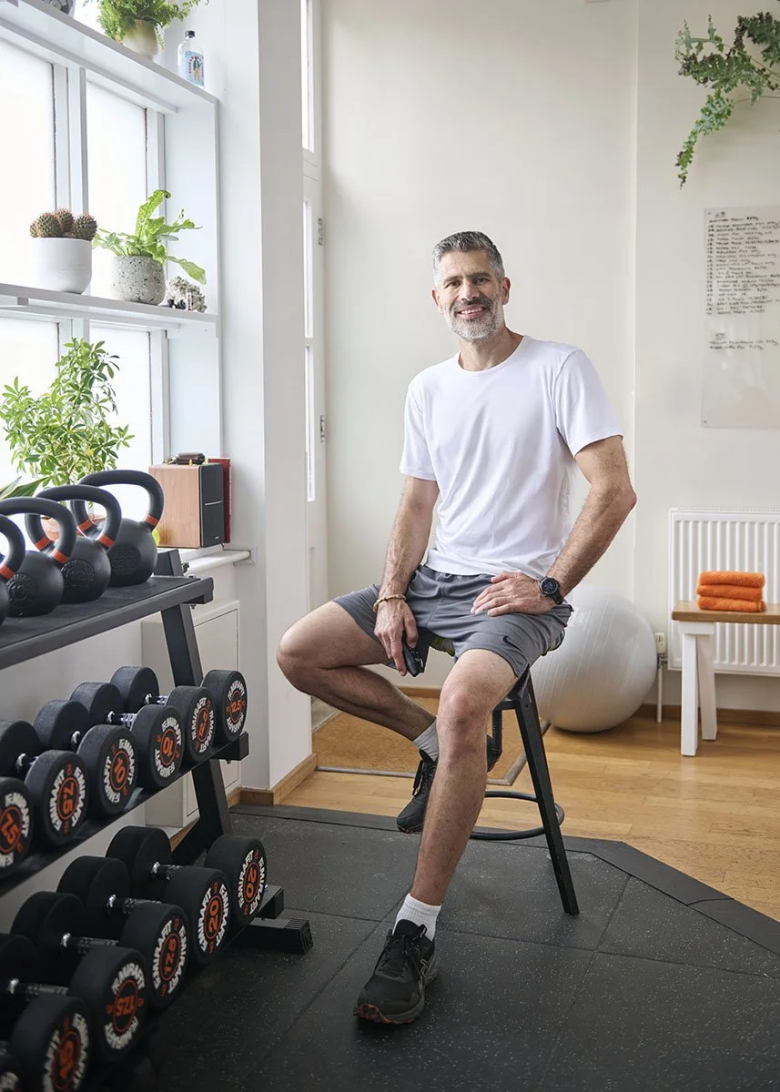 A middle-aged man with gray hair and a beard, wearing a white t-shirt, gray shorts, and black athletic shoes, sitting on a stool in a home gym. The gym has kettlebells and dumbbells on a rack, with potted plants and a large exercise ball in the background.