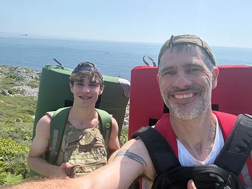 A man and a girl taking a selfie outdoors near the ocean with backpacks, smiling.