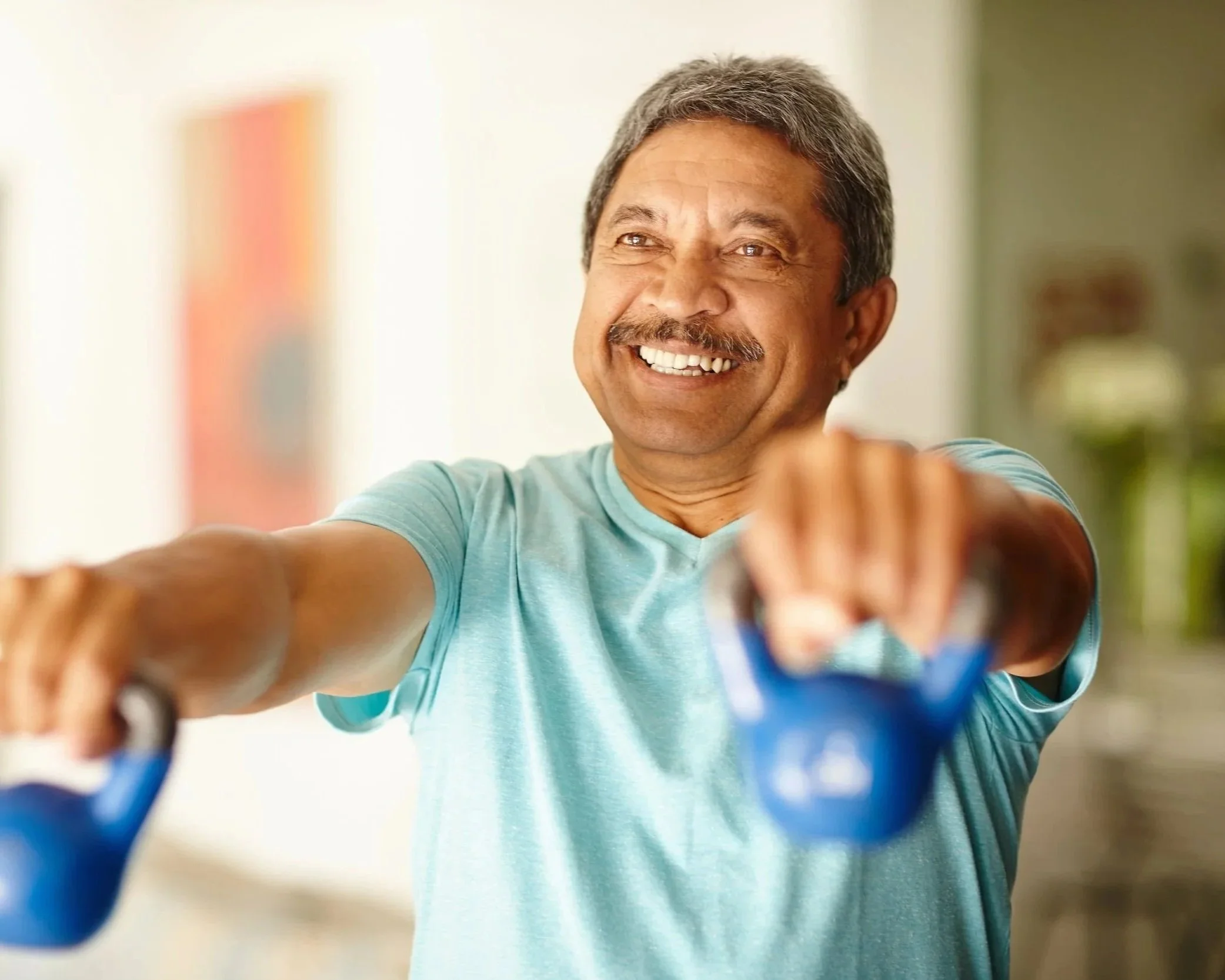 Close-up of a middle-aged man with gray hair and a mustache smiling and reaching out his hand, with children in the foreground and a blurred indoor background.  Personal Training for the over 50's. Ramsbury, Wiltshire.