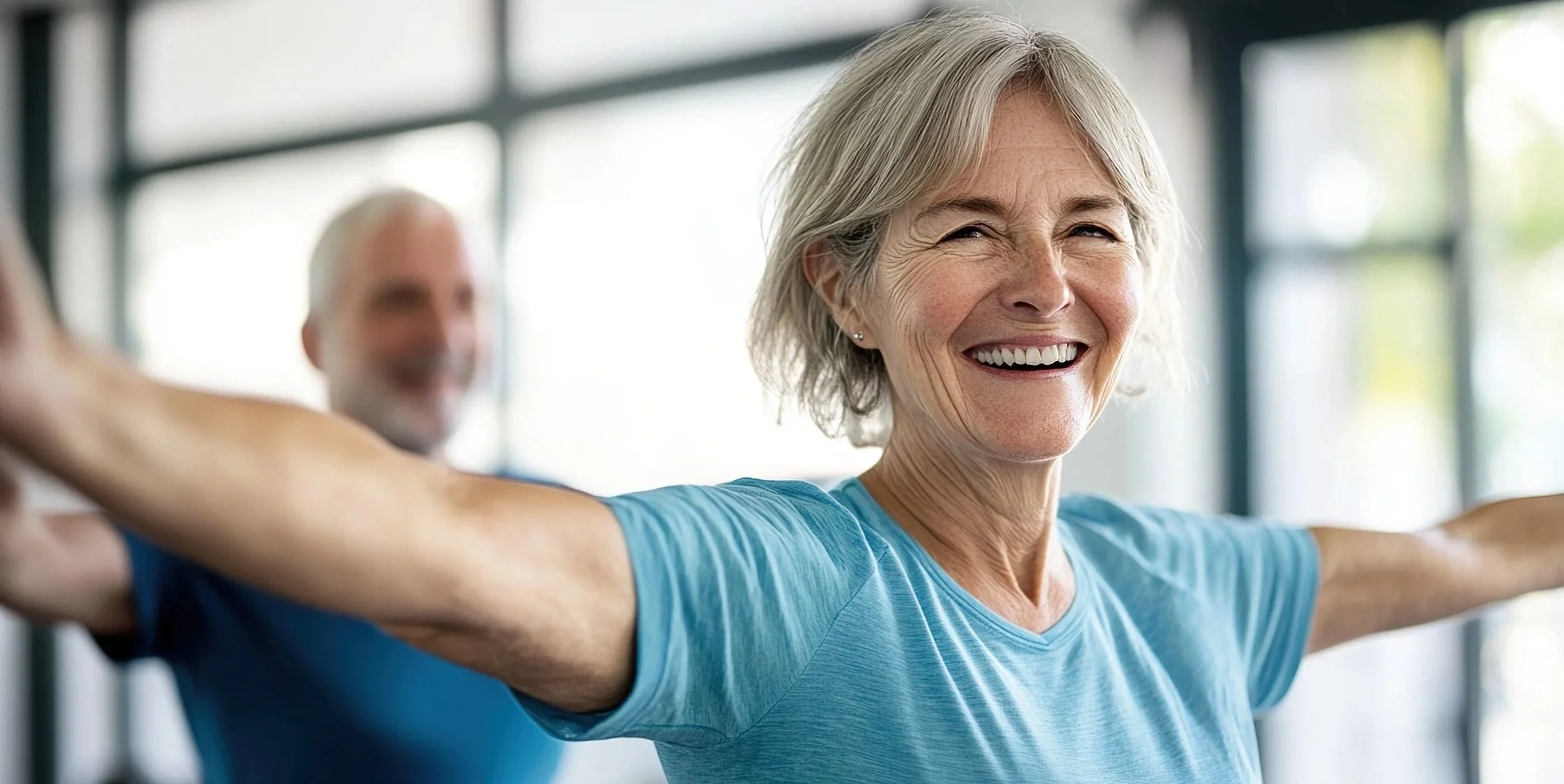 Senior woman with gray hair smiling during a workout class, stretching arms in a bright indoor gym. Personal Training for the over 50's. Ramsbury, Wiltshire.