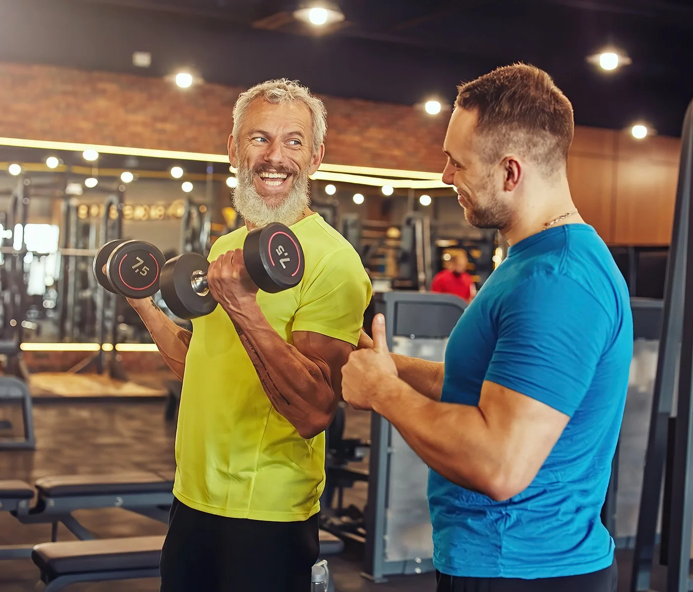 Two men in a gym, one lifting a dumbbell and the other giving a thumbs up, both smiling and engaging in conversation.  Personal Training for the over 50's. Ramsbury, Wiltshire.
