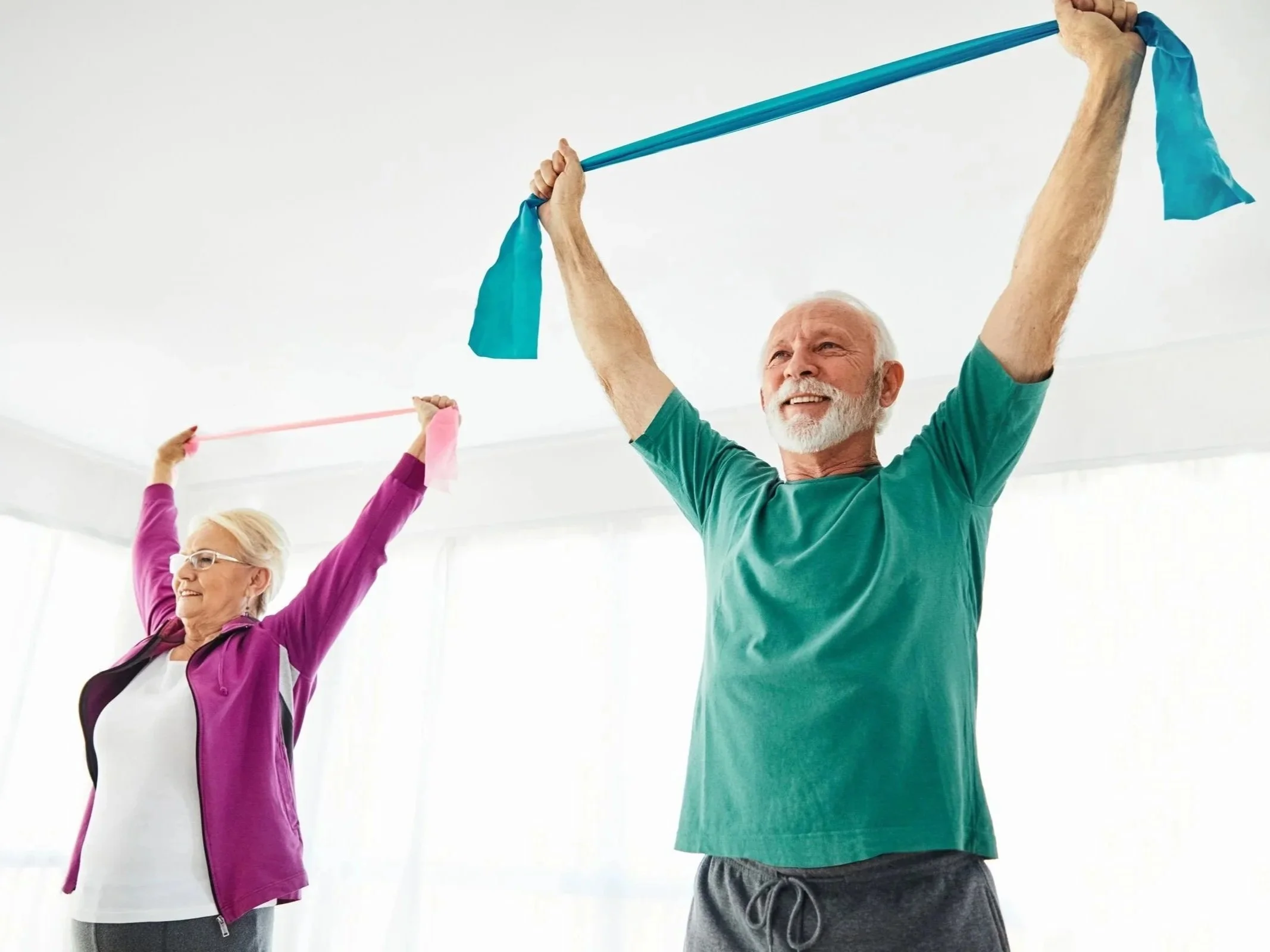 An elderly woman and a younger man are practicing yoga or stretching exercises indoors. Personal Training for the over 50's. Ramsbury, Wiltshire.