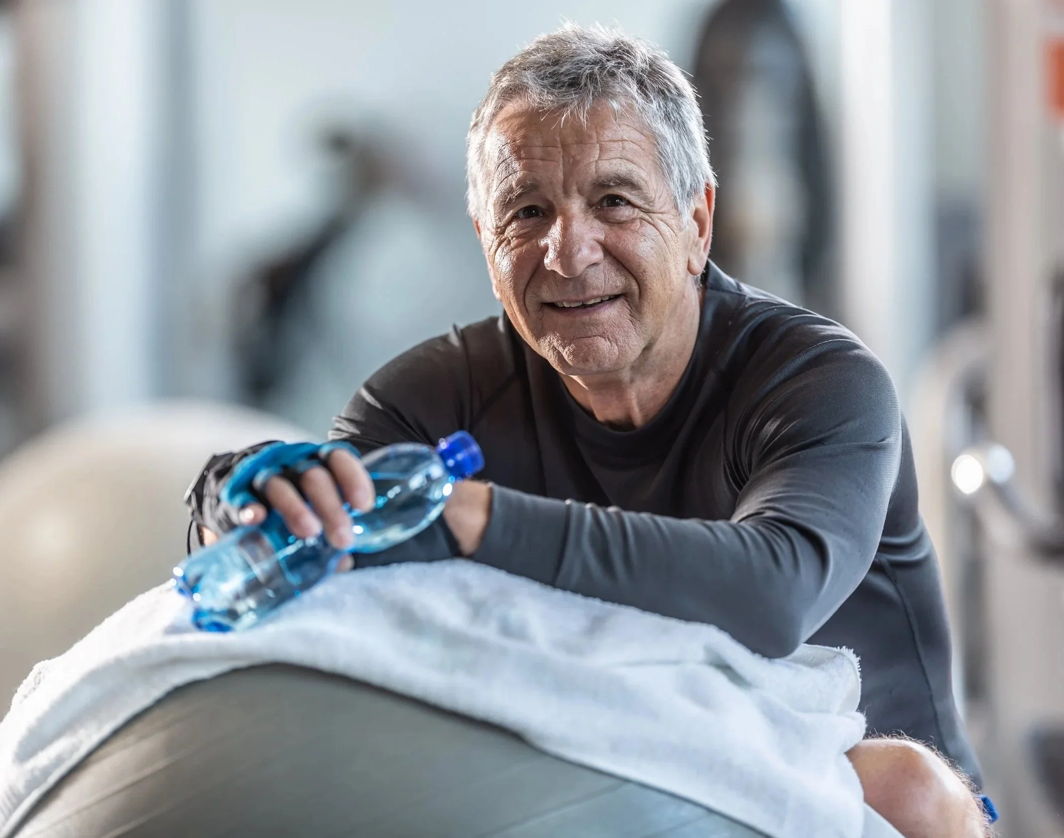 Smiling senior man in athletic wear enjoying a workout at gym, bending over a mat with a water bottle, with a blurred gym background.  Personal Training for the over 50's. Ramsbury, Wiltshire.