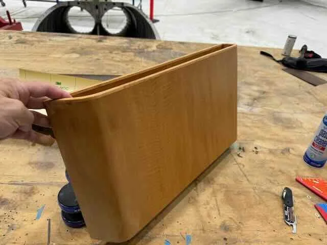 A person holding a wooden bookend or magazine holder on a workbench, with tools and a bottle of glue nearby in a workshop or garage setting.