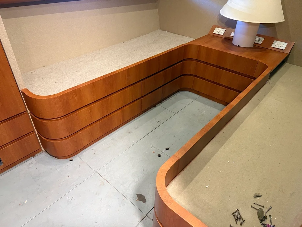 A wooden bed frame and headboard with power outlets and a lamp at the head of the bed, missing mattress, in a hotel room.