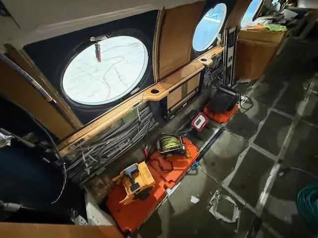 Inside a bus or ship with a wooden interior, tools and equipment on an orange workbench, wiring along the walls, and large oval windows.
